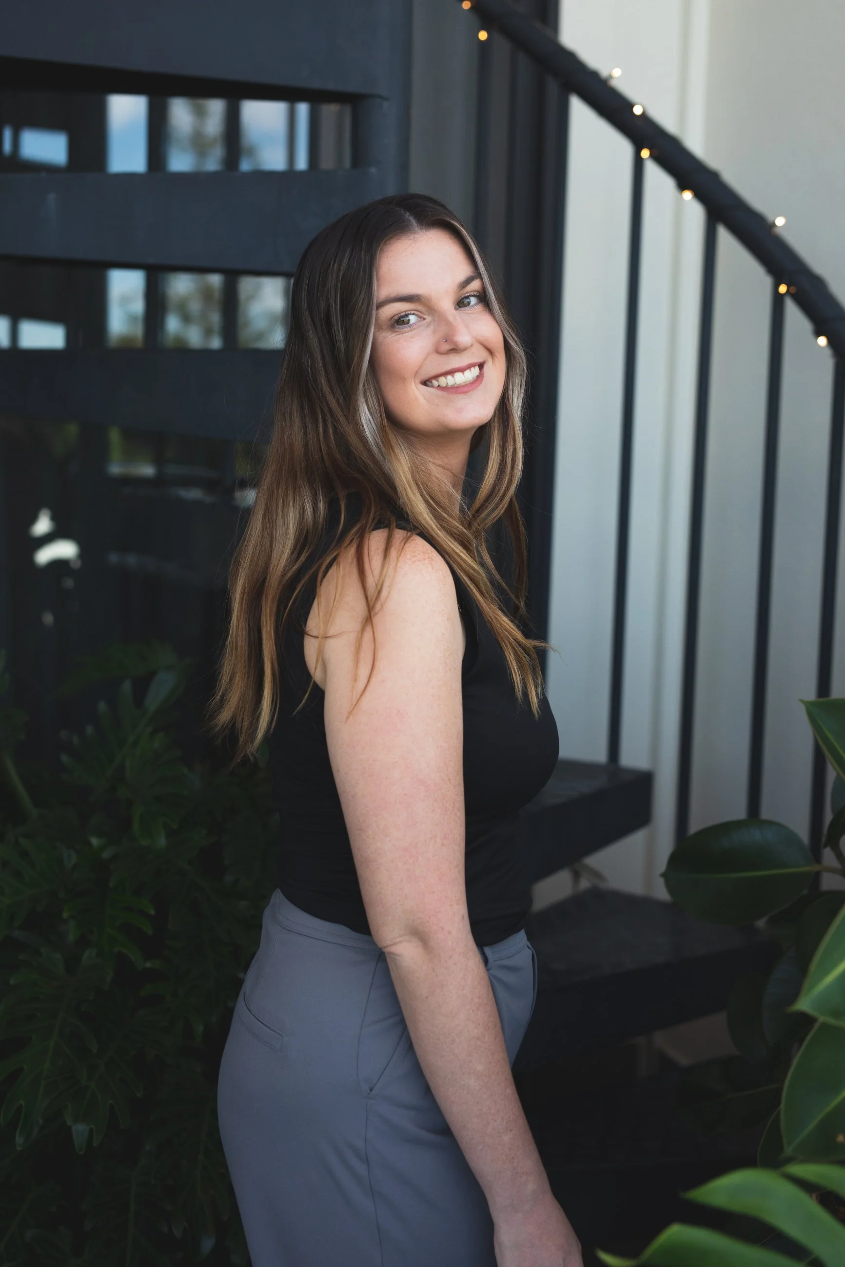 A woman with long brown hair smiling, wearing a black sleeveless top and gray pants, standing next to a black staircase with string lights and green plants nearby.