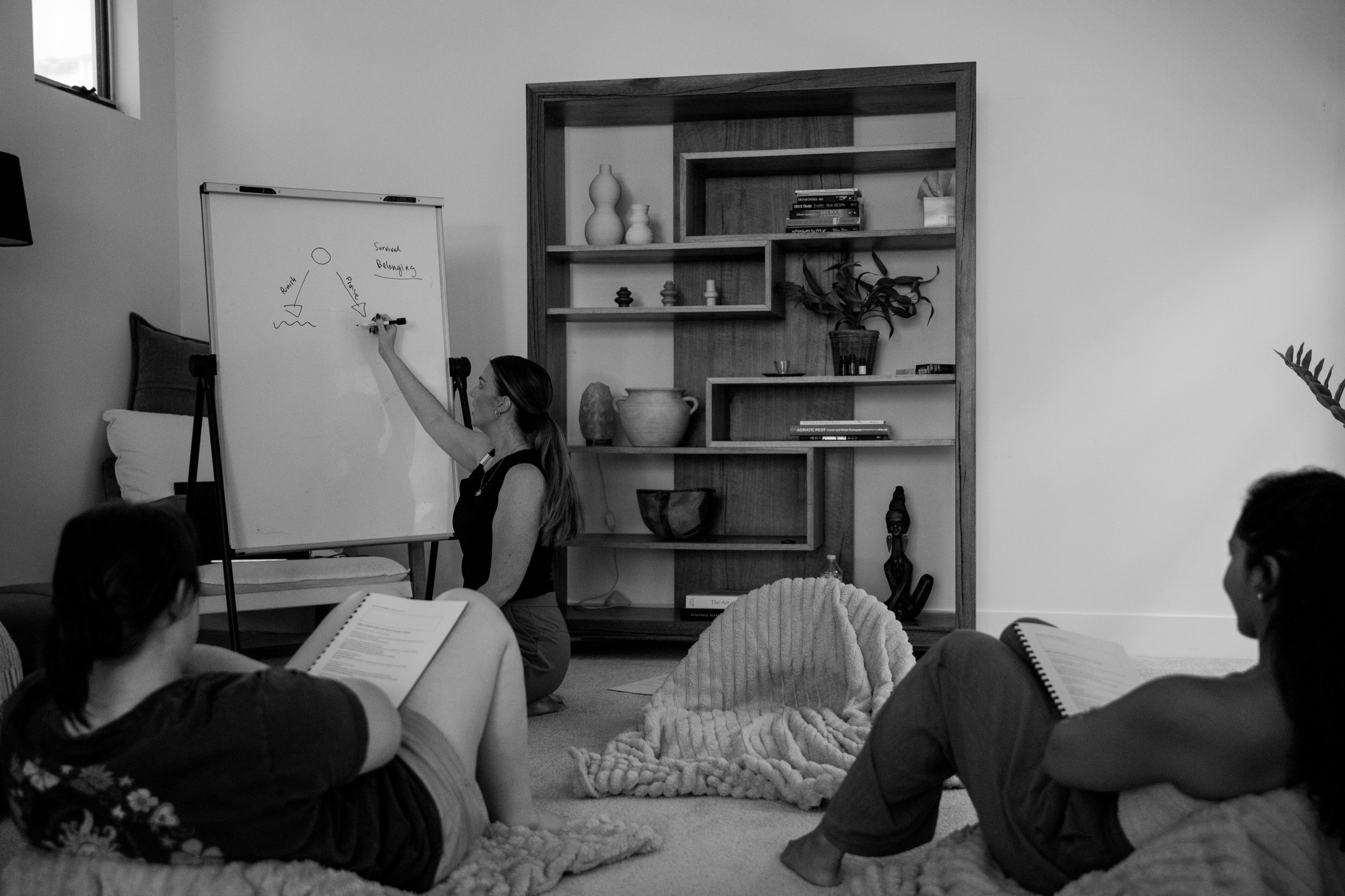 A woman is standing and pointing at a whiteboard with diagrams inside a room, while several children are sitting on the floor, some reading books and others listening to her.