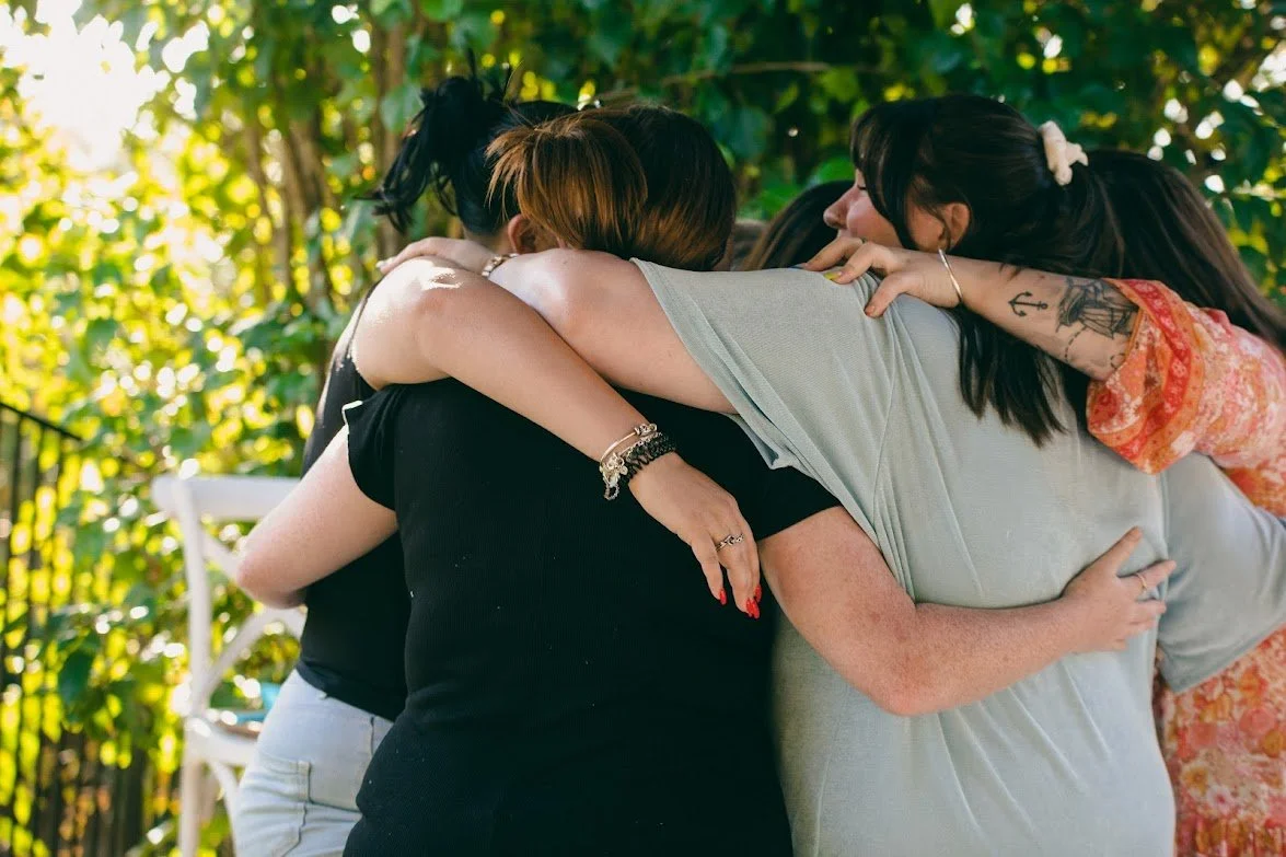 A group of women hugging outdoors in a circle, surrounded by greenery and sunlight.