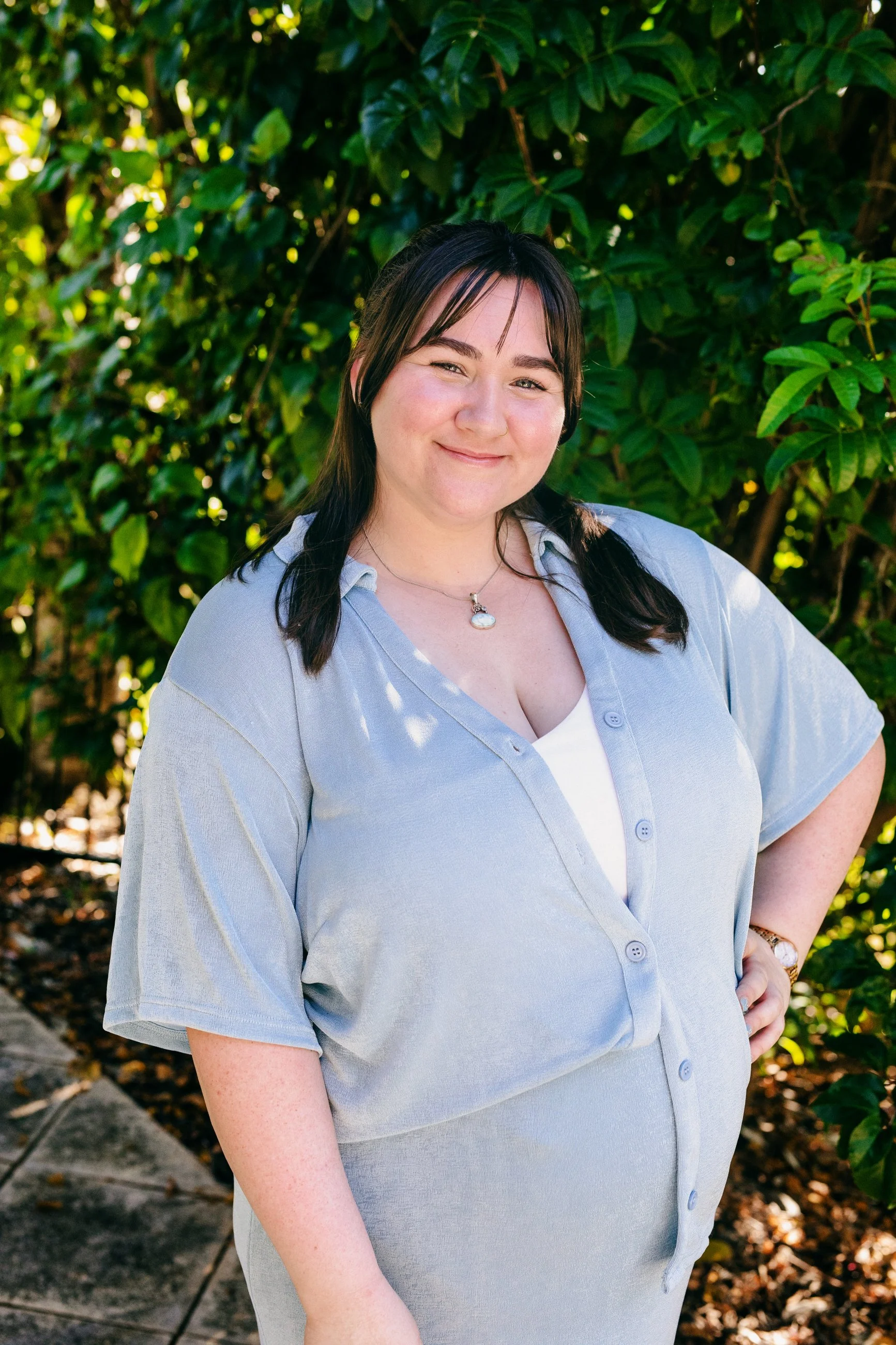 A woman with shoulder-length dark brown hair, smiling, standing outdoors in front of green foliage. She is wearing a light blue button-up shirt over a white top, accessorized with a necklace and a watch, with one hand on her hip.