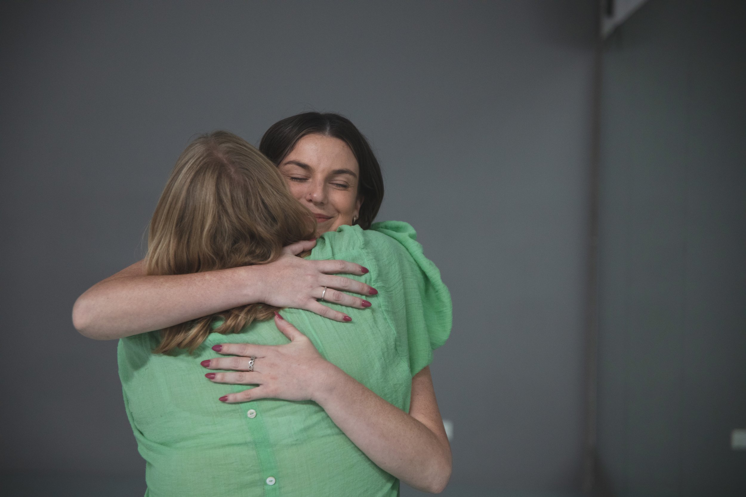 Two women hugging in a close embrace, smiling. One woman has long blonde hair, and the other has short black hair and is wearing a green top.
