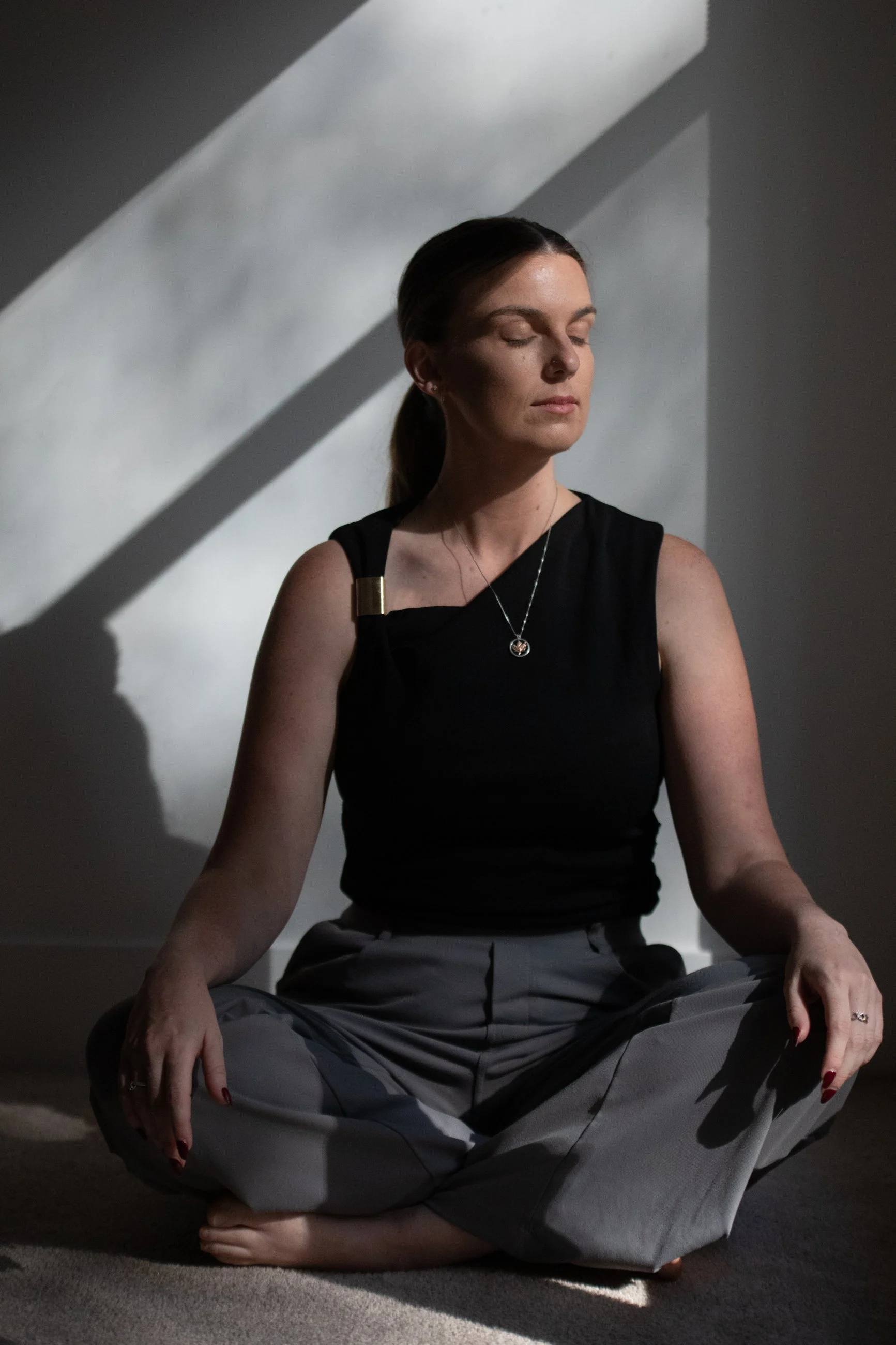 A woman practicing yoga or meditation in a seated cross-legged pose with her eyes closed, sitting on the floor in a dimly lit room with shadows cast on the wall behind her.