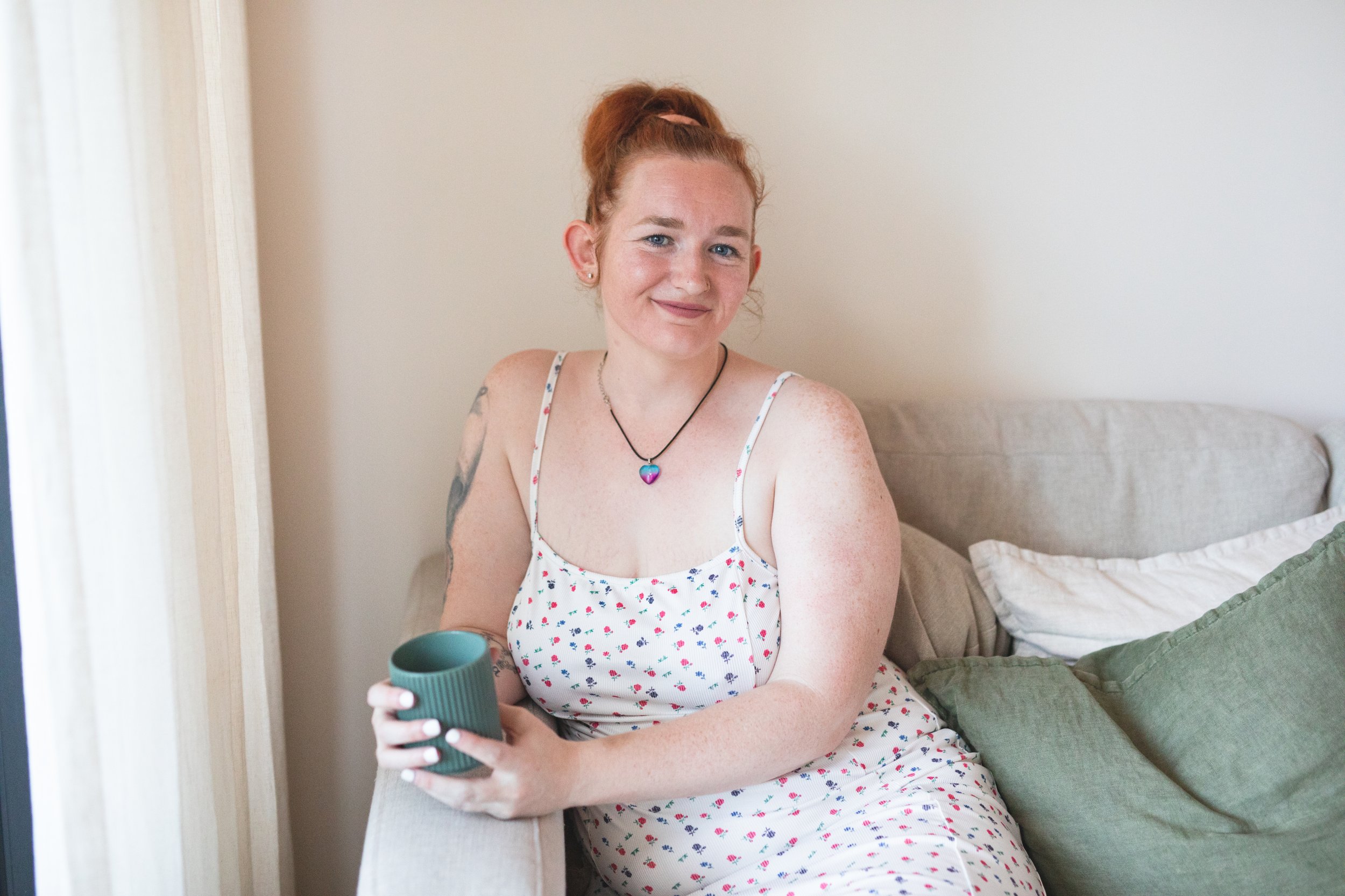 A young woman with red hair, blue eyes, and freckles, smiling, sitting on a bed holding a dark green cup, wearing a white dress with colorful flower prints and a heart-shaped pendant necklace.