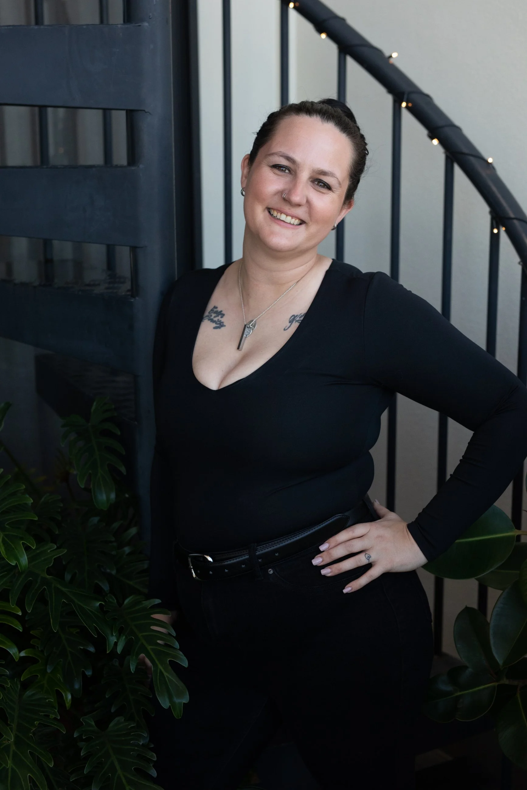 A woman with dark hair in a bun, wearing a black long-sleeve top and black pants, stands outdoors next to a black staircase and plants, smiling at the camera.