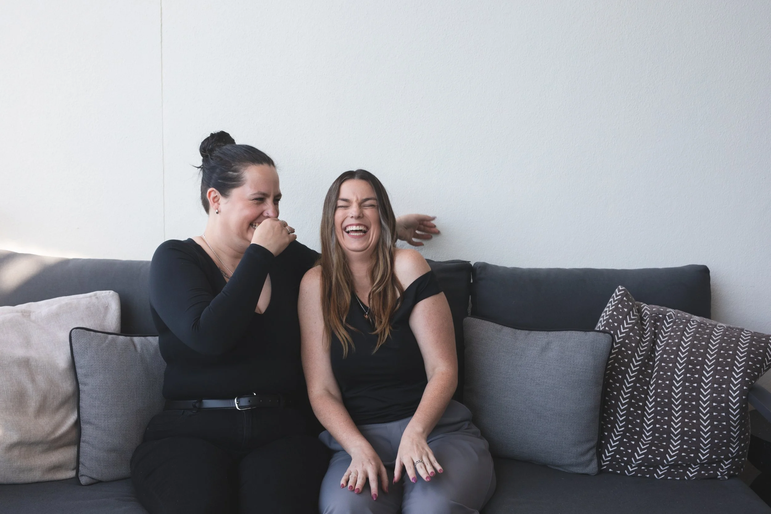 Two women sitting on a couch sharing a laugh, with a plain wall behind them and multiple cushions on the sofa.