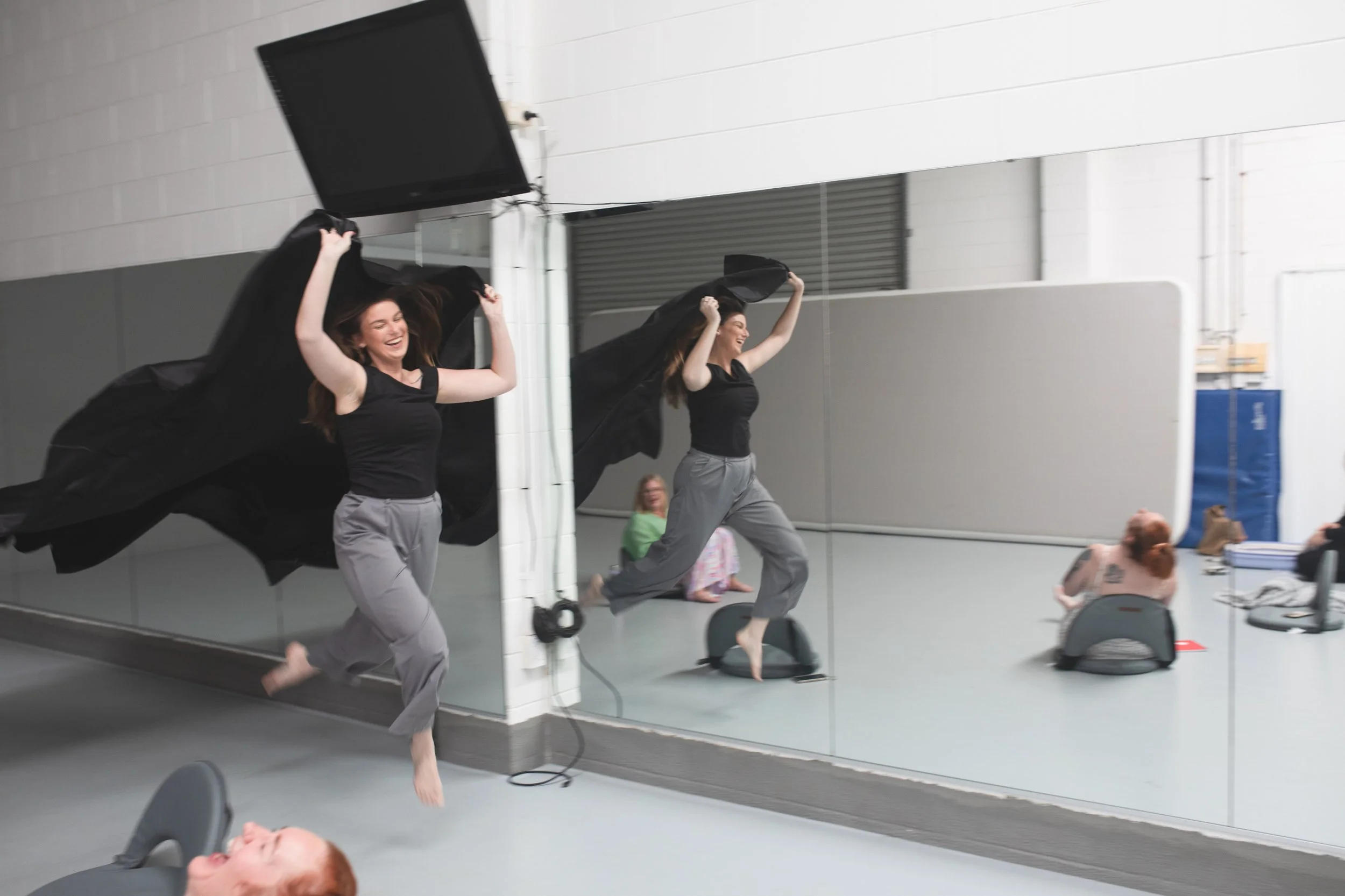 Two women in black tops and gray pants are joyfully running in front of a mirror, with black fabric flowing behind them, in a room with white walls and a gray floor. Several children and adults are seated or lying on the floor and chairs nearby.