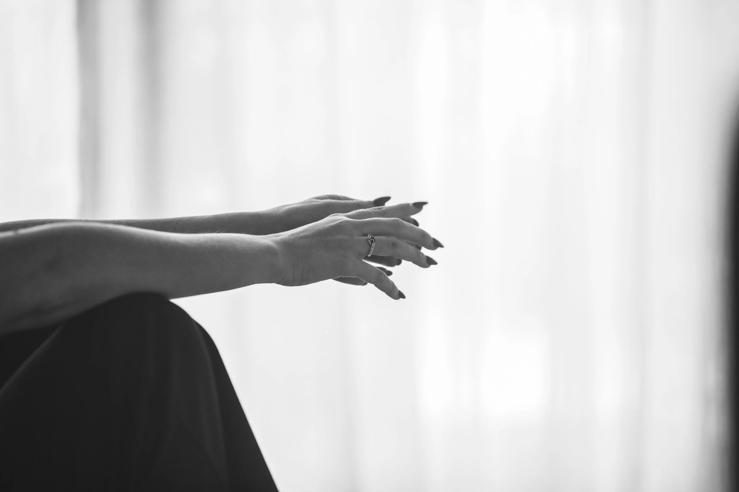 Black and white photo of a person's outstretched hands and arms resting on their crossed knee, with a ring on one finger, against a blurred light-colored background.