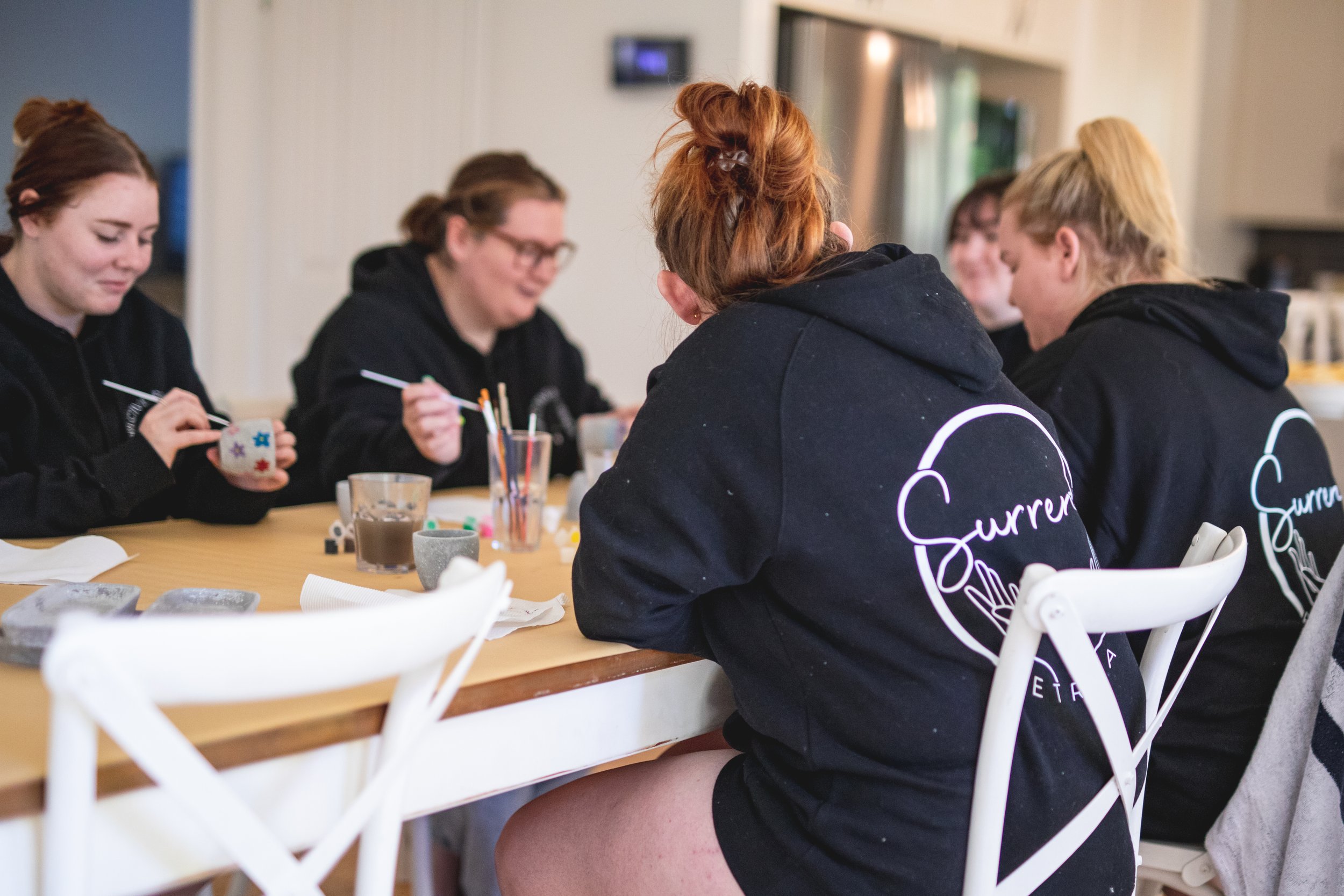 Group of young women gathered around a table, participating in arts and crafts activity, with craft supplies and beverages on the table.