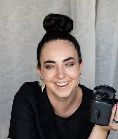 A woman with dark hair in a high bun, smiling, wearing earrings, holding a camera, and sitting in front of a curtain.