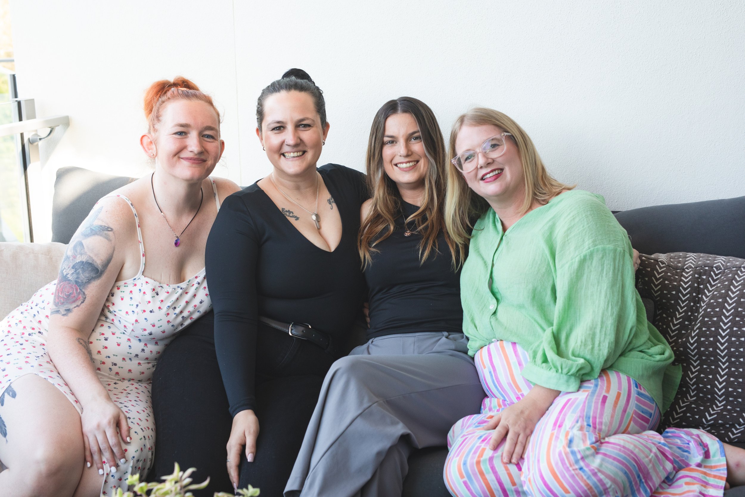 Four women sitting together on a couch, smiling at the camera in a well-lit room.
