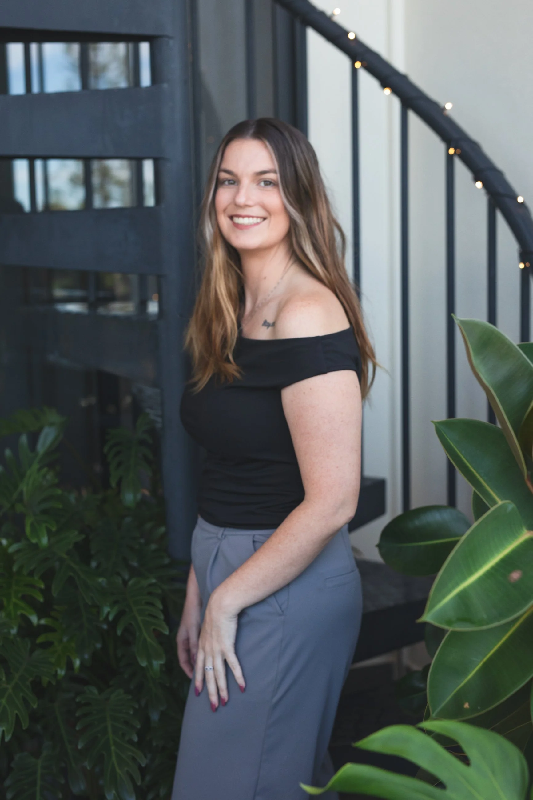 A young woman with long, wavy brown hair, wearing an off-the-shoulder black top and gray pants, smiling while standing next to greenery and a staircase railing.