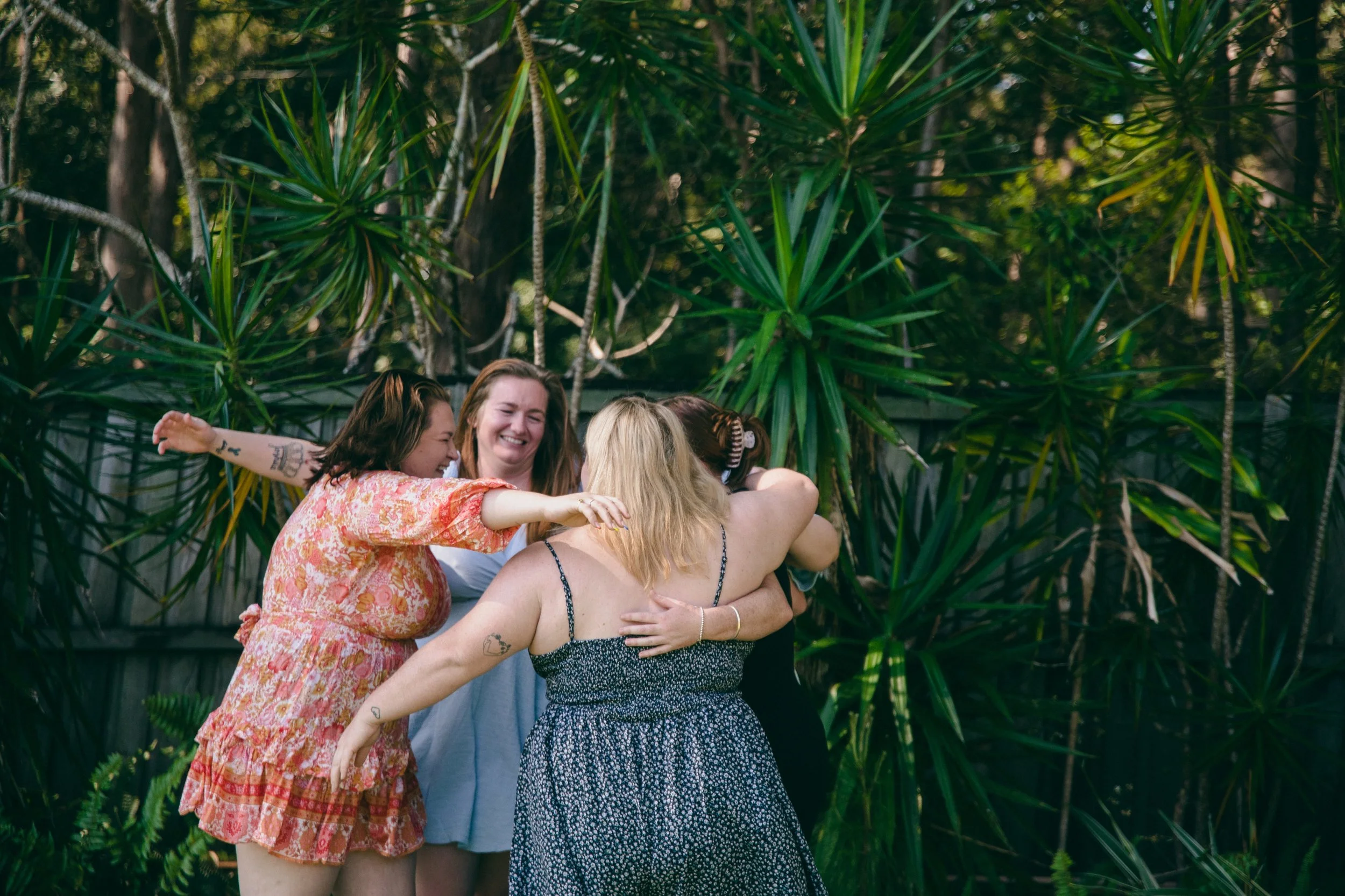 Group of women hugging and smiling outdoors surrounded by lush green plants and trees.