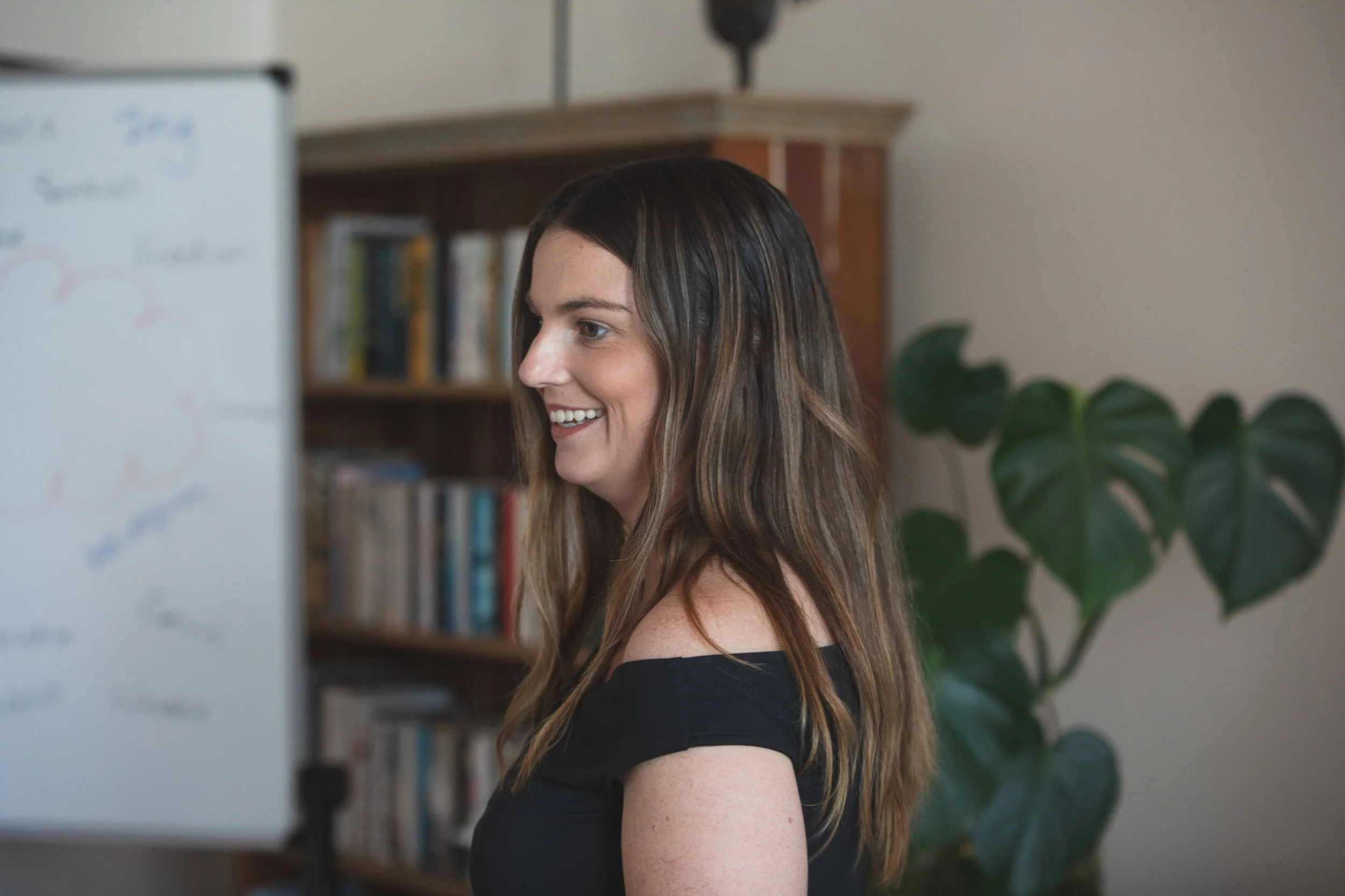 A woman with long brown hair smiling while looking at a whiteboard in a room with a bookshelf and houseplant.