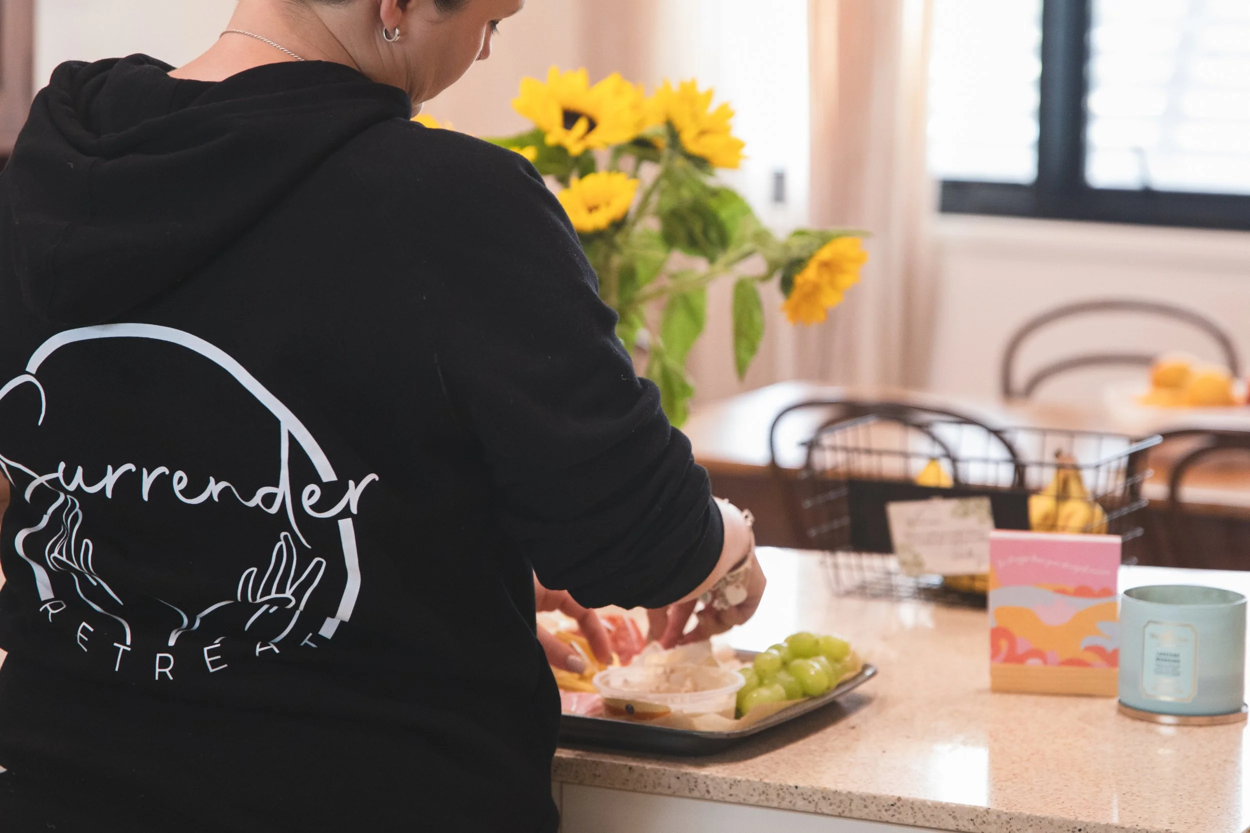 Person wearing a black hoodie preparing food with grapes and other items on a kitchen countertop, with sunflowers in the background.
