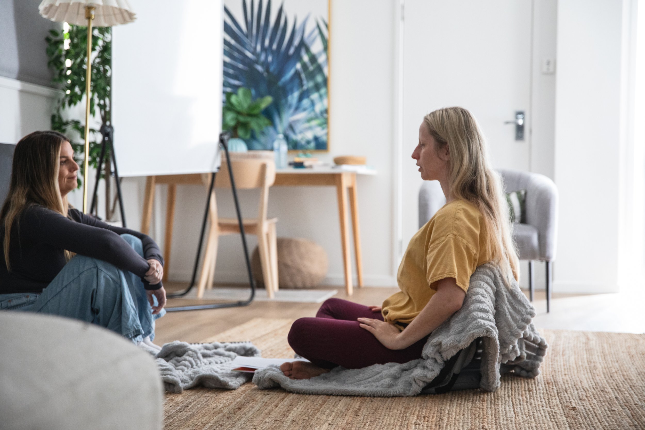 Two women sitting cross-legged on a rug, meditating in a living room.