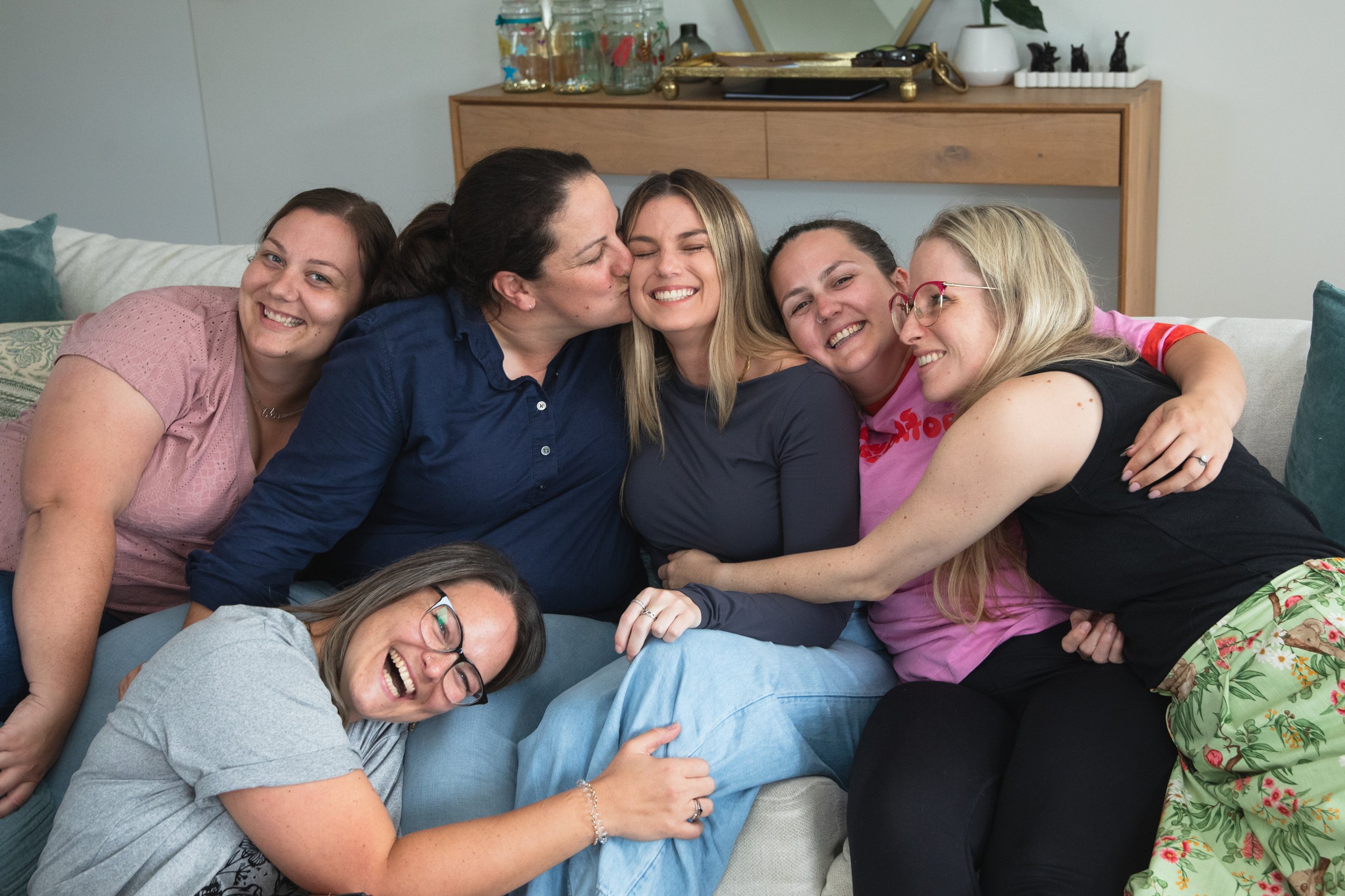 Group of six women hugging and smiling on a couch in a living room.