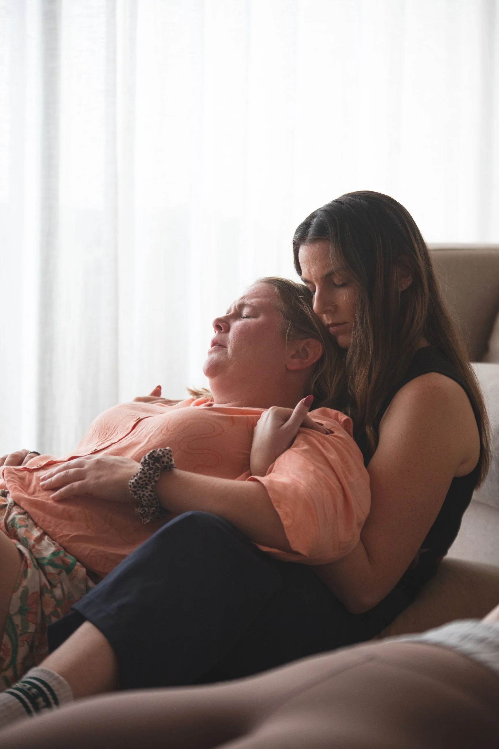 A woman with long dark hair holds an elderly woman with short blond hair and closed eyes in a comforting embrace indoors near a window with white curtains.