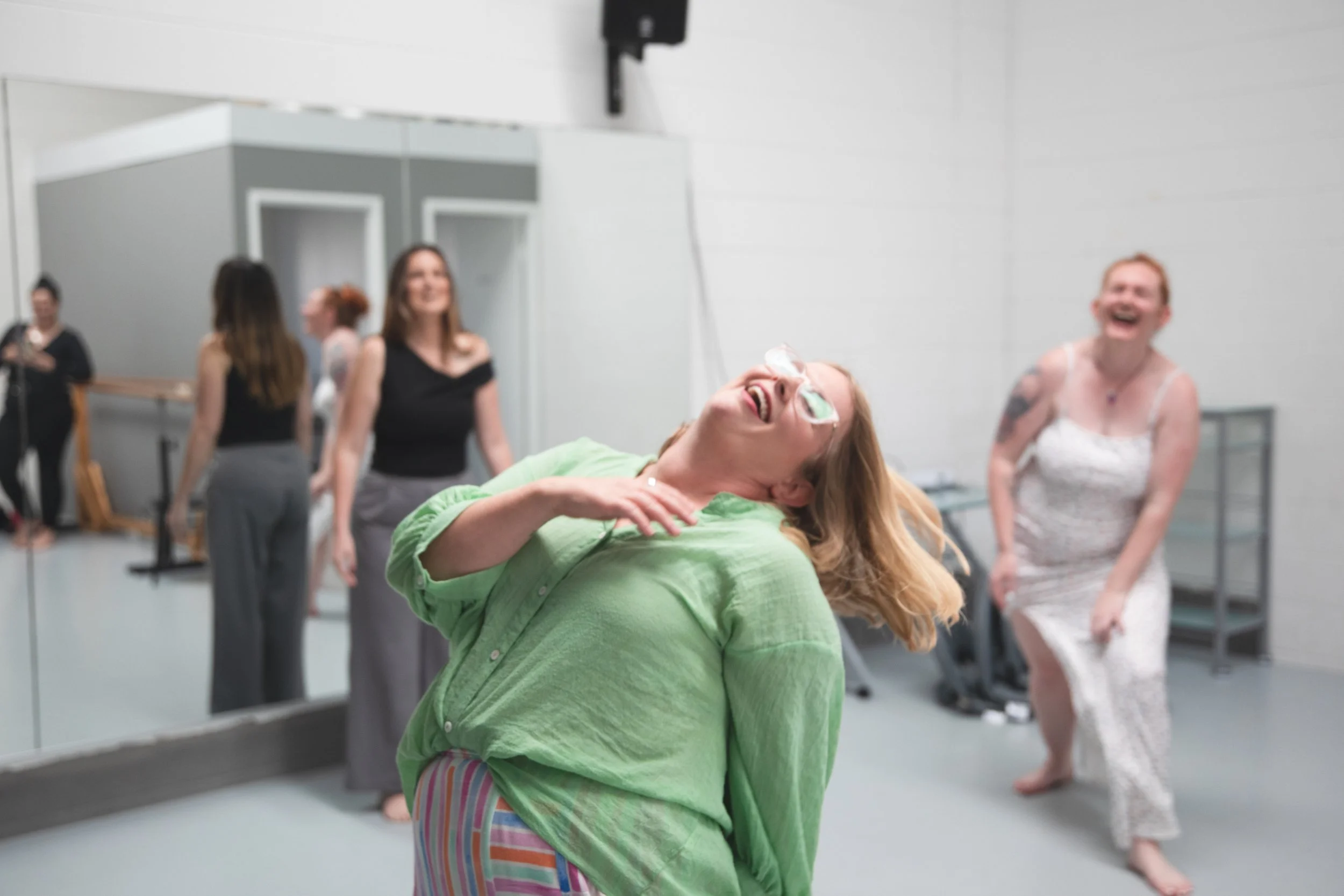 A young woman in a green shirt and striped pants laughing and leaning back with her head tilted, while other women in dress and casual clothing are standing and sitting in a room with white walls and large mirrors, likely a dance or art studio.