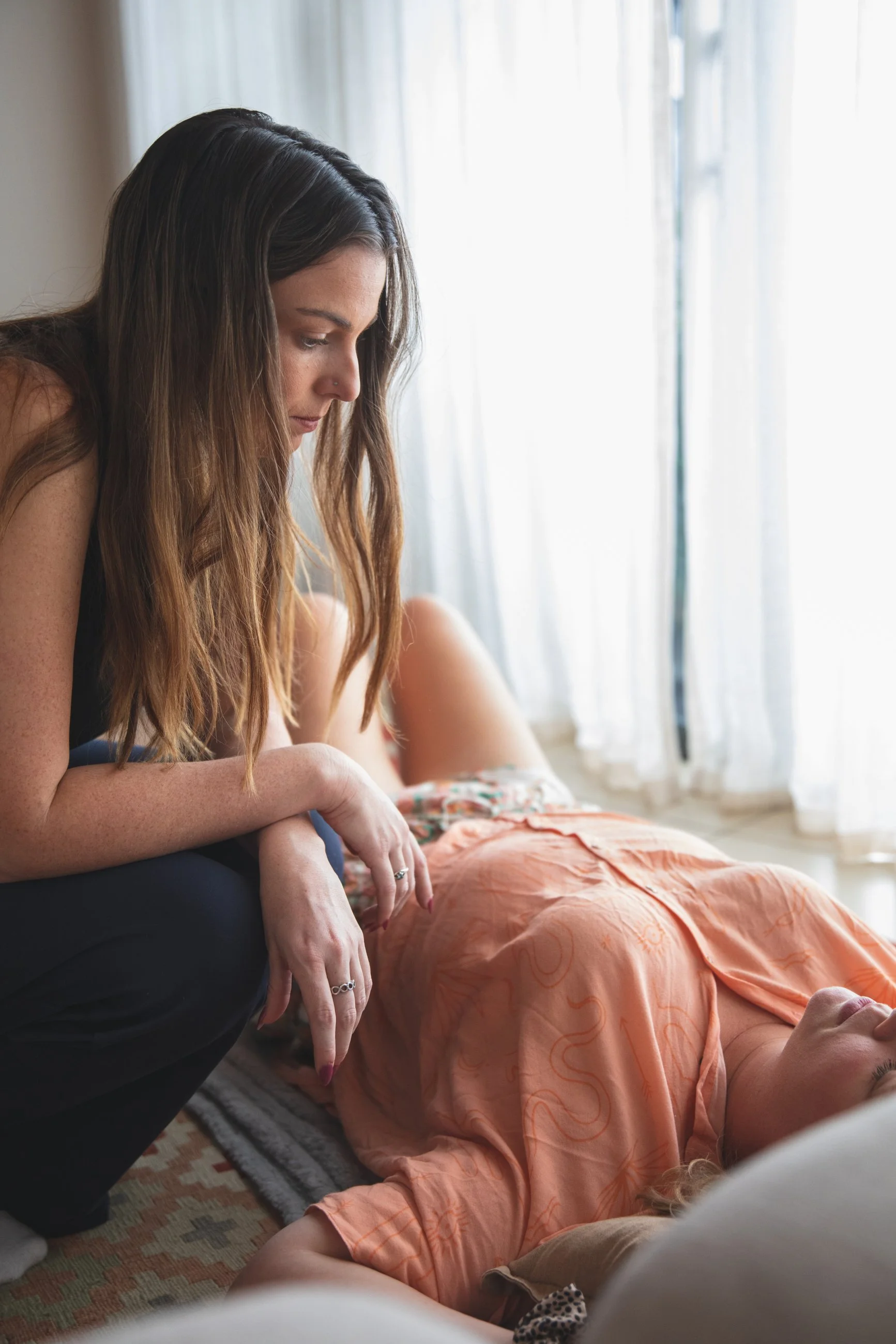 A woman with long brown hair crouches beside a woman lying on the floor, looking at her intently. The woman on the floor has blonde hair and is wearing an orange shirt, lying on a rug near a window with sheer curtains.
