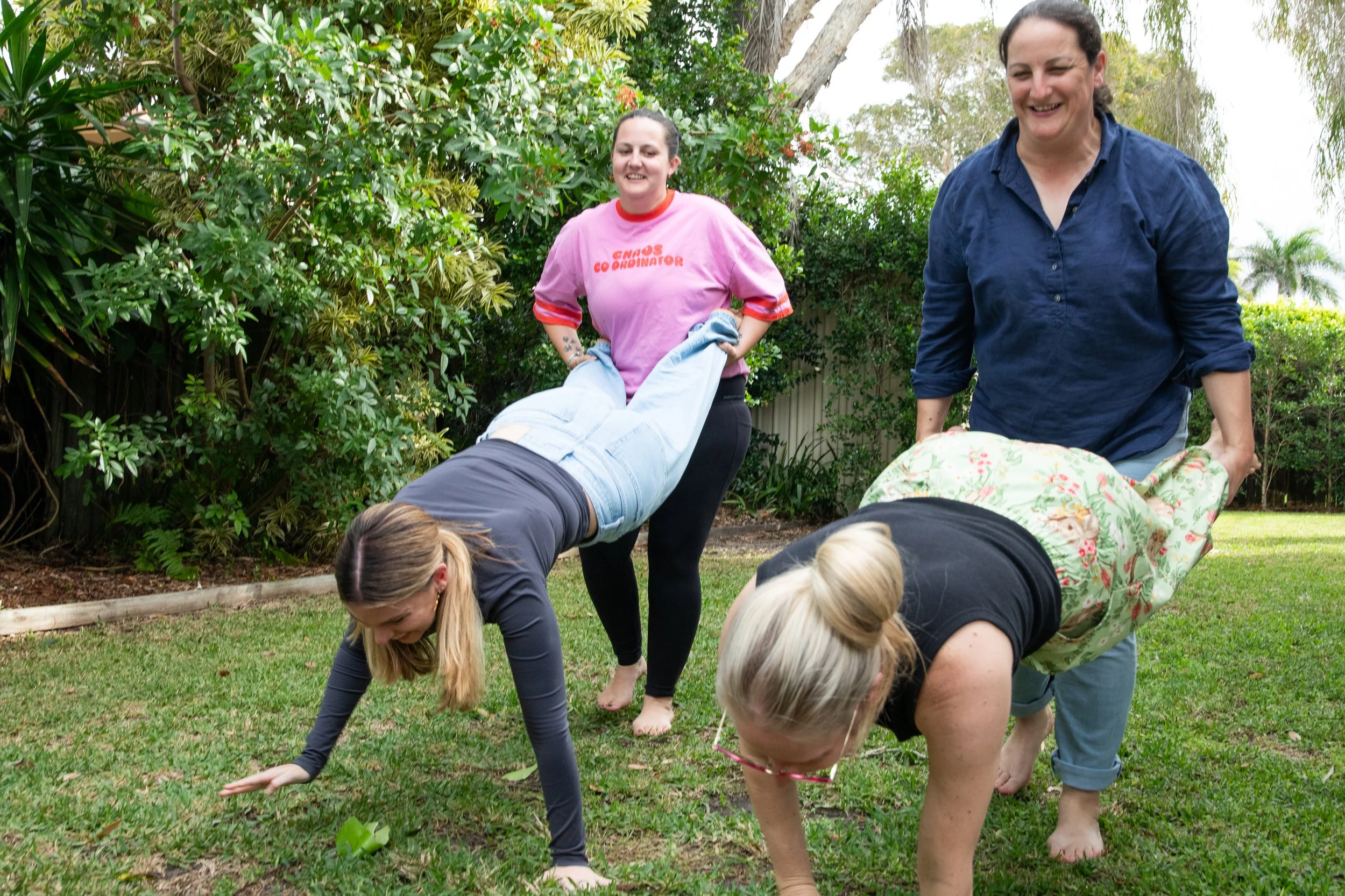 People participating in outdoor yoga class, bending forward on grass, surrounded by lush greenery and trees.