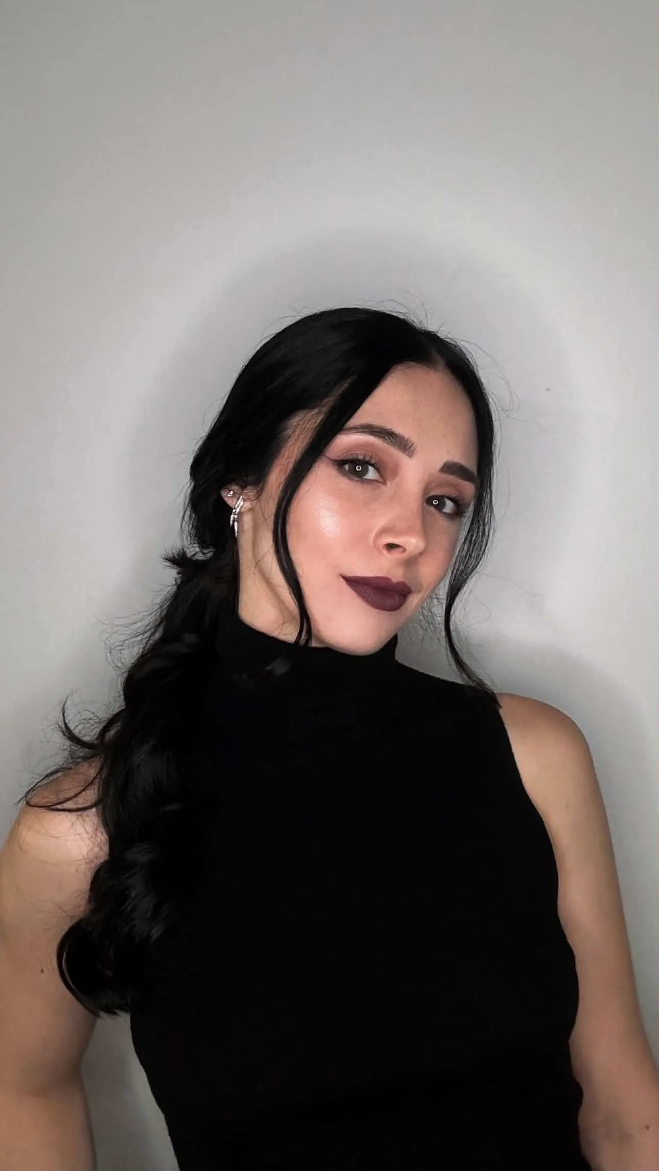 A woman with long dark hair and dark lipstick taking a selfie against a plain light gray background. She is wearing a black sleeveless top and silver earrings.