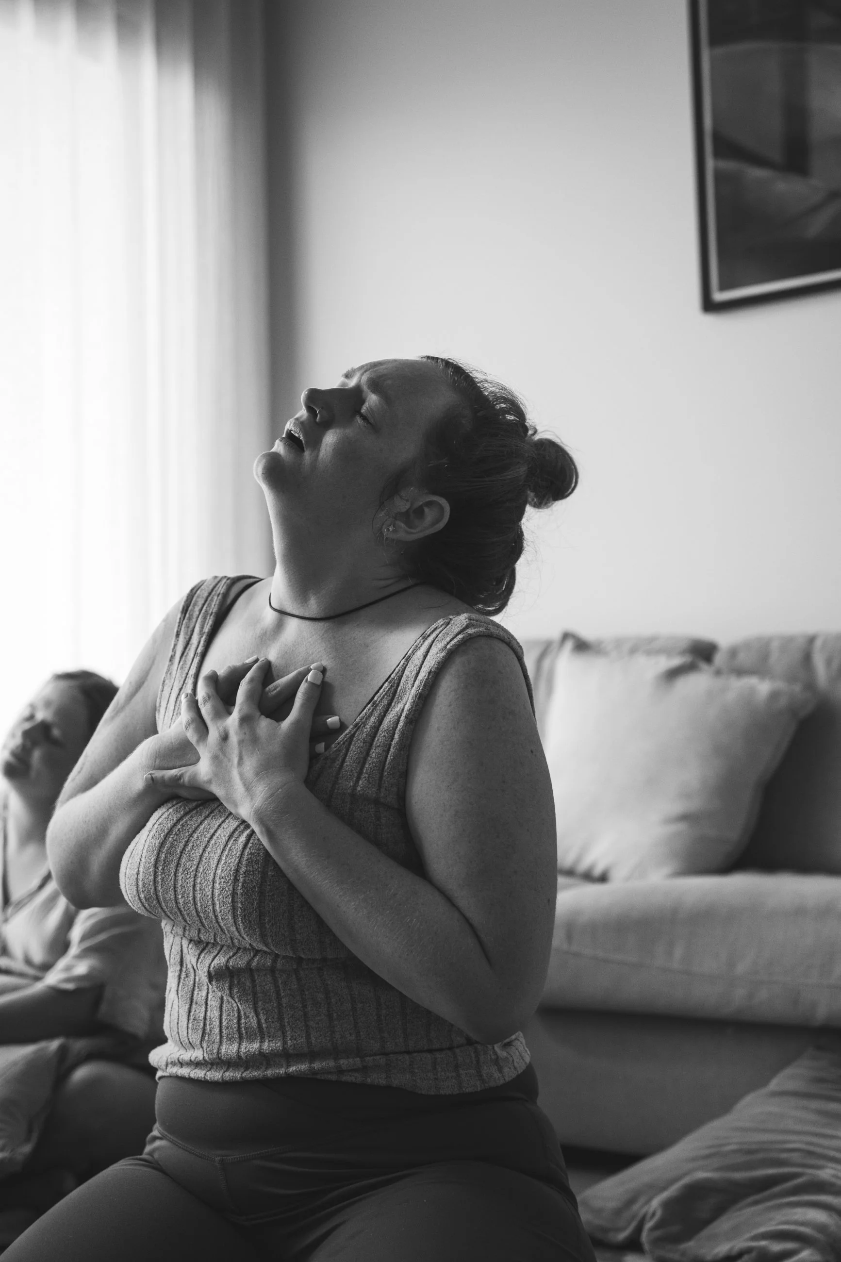 A woman is kneeling on the floor with her hand on her chest and appears to be experiencing a moment of intense emotion or pain inside a living room with a sofa and a framed picture on the wall.
