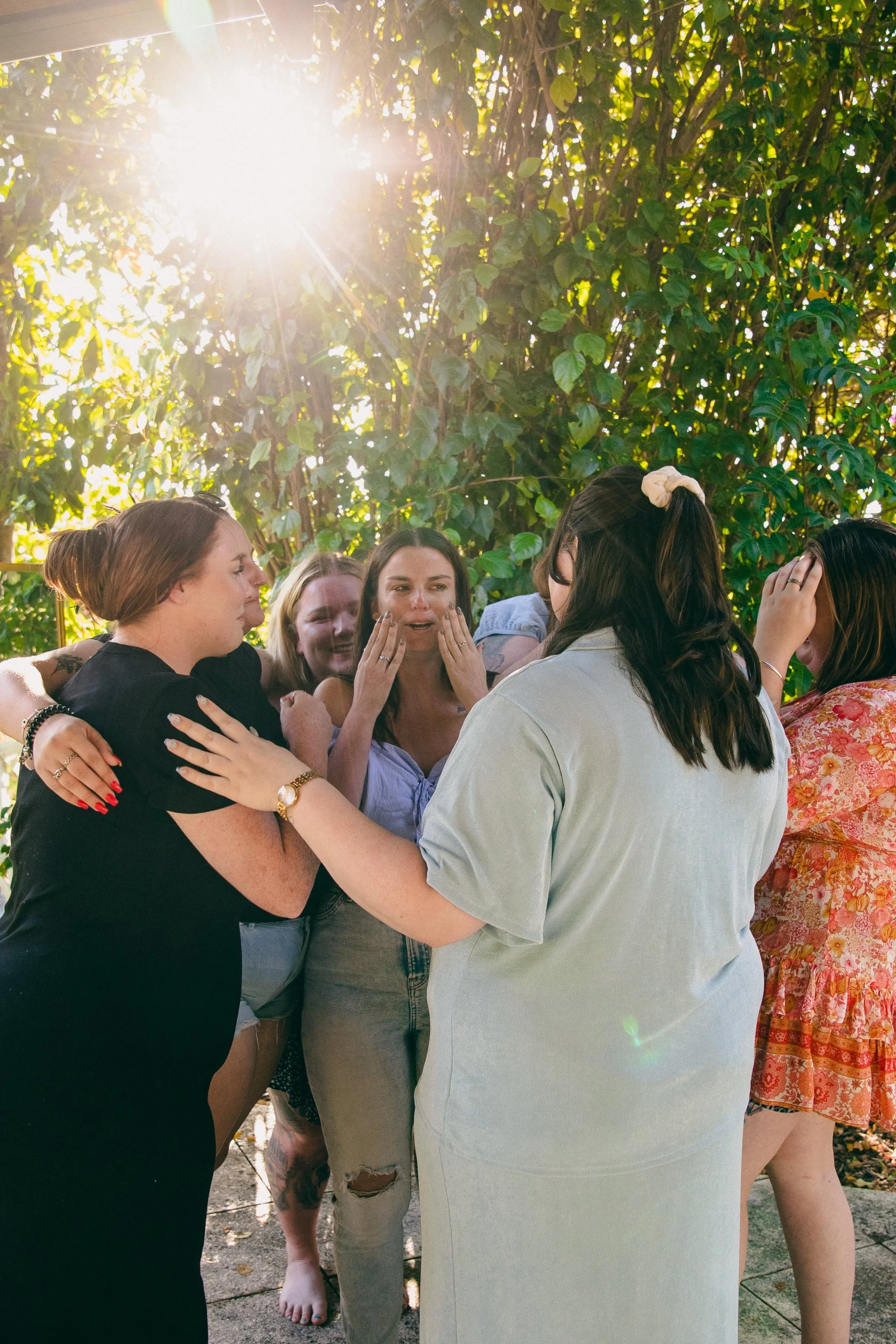 A group of women are gathered outdoors, embracing and comforting each other under the sunlight with greenery in the background.