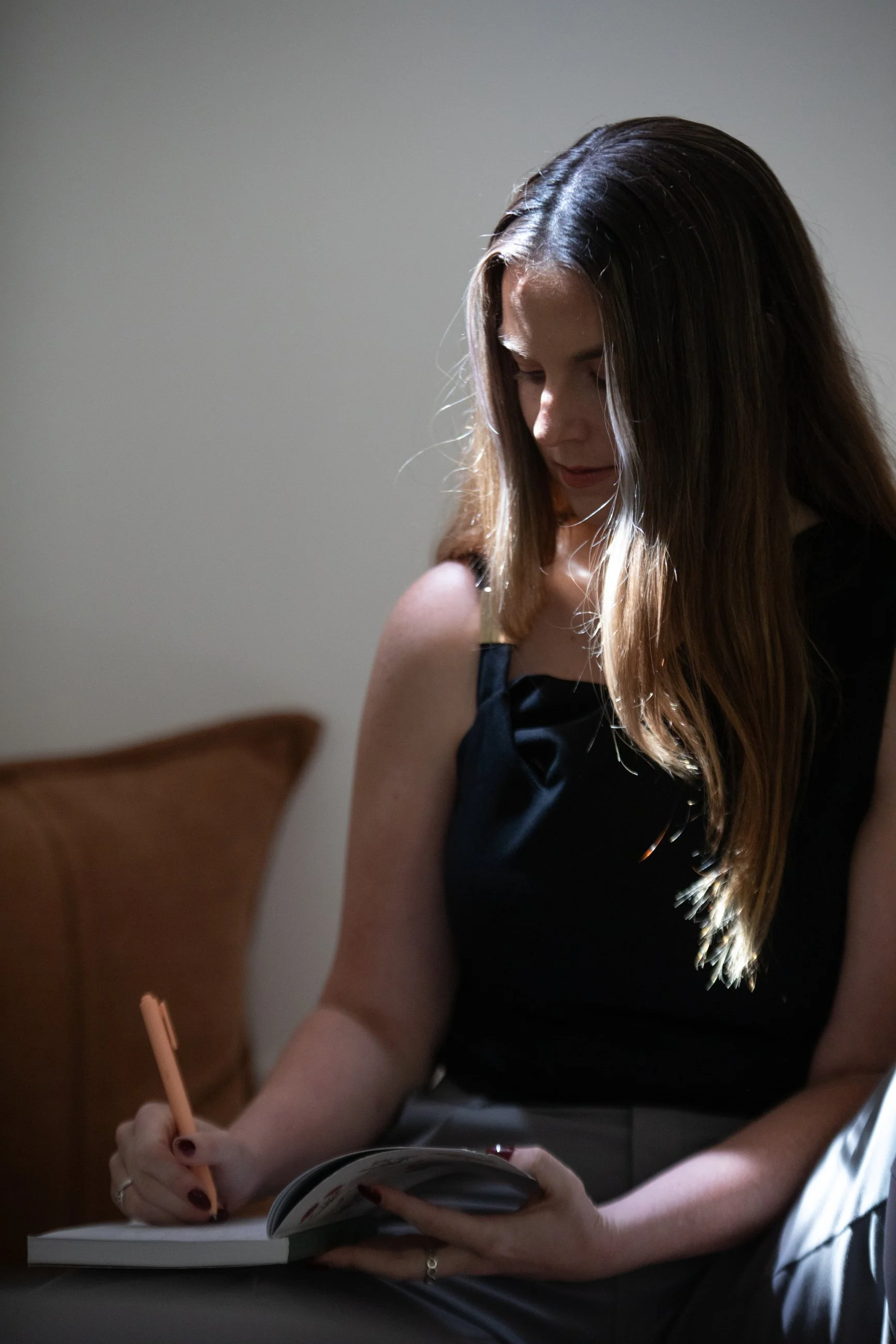 A woman with long brown hair wearing a black sleeveless top is sitting at a table, writing or drawing in a notebook with a light-colored pen. She is looking downward at her notebook, and her right hand is holding the pen.