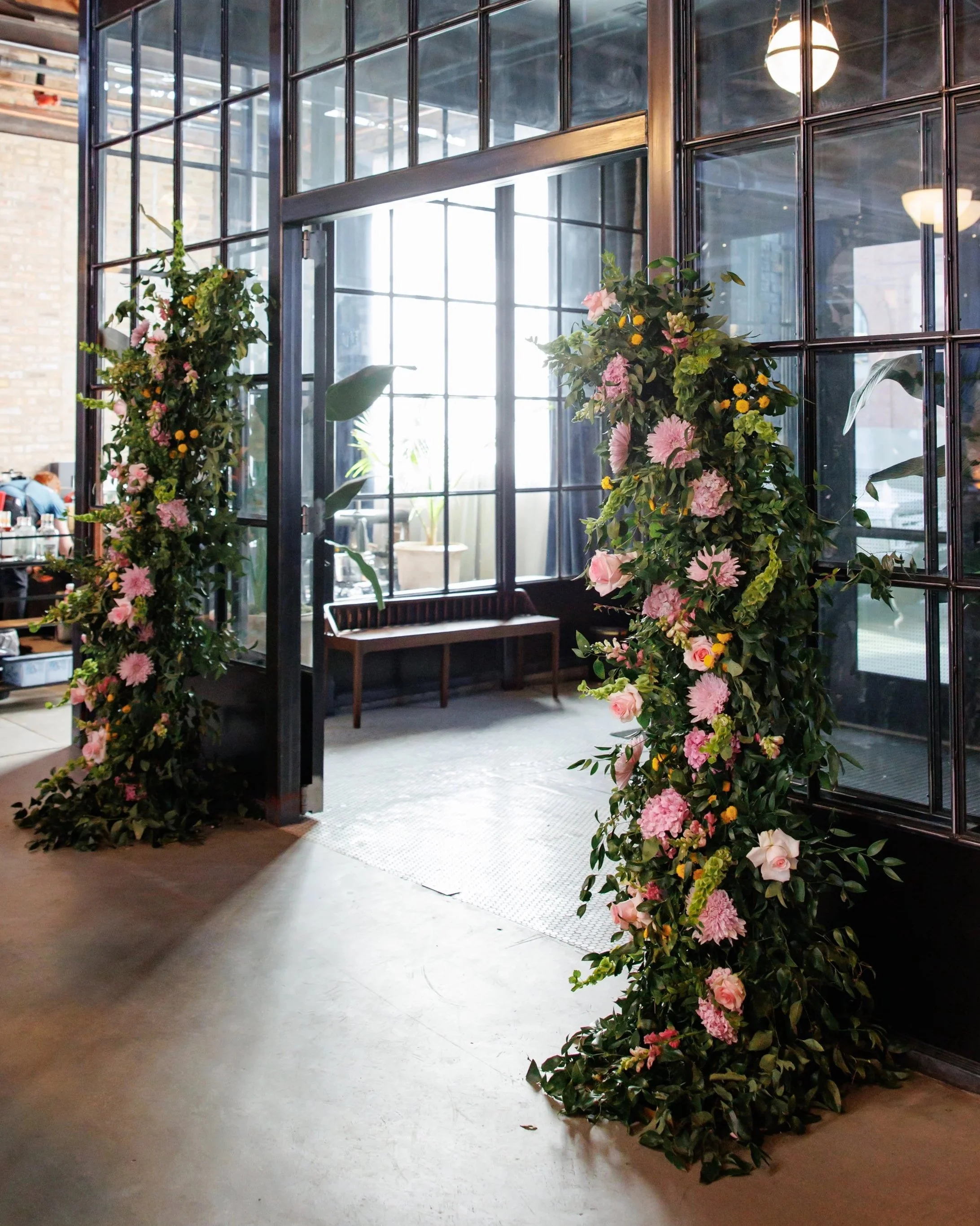 Decorated floral archway at the entrance of a modern industrial-style venue with glass walls and a bench inside.