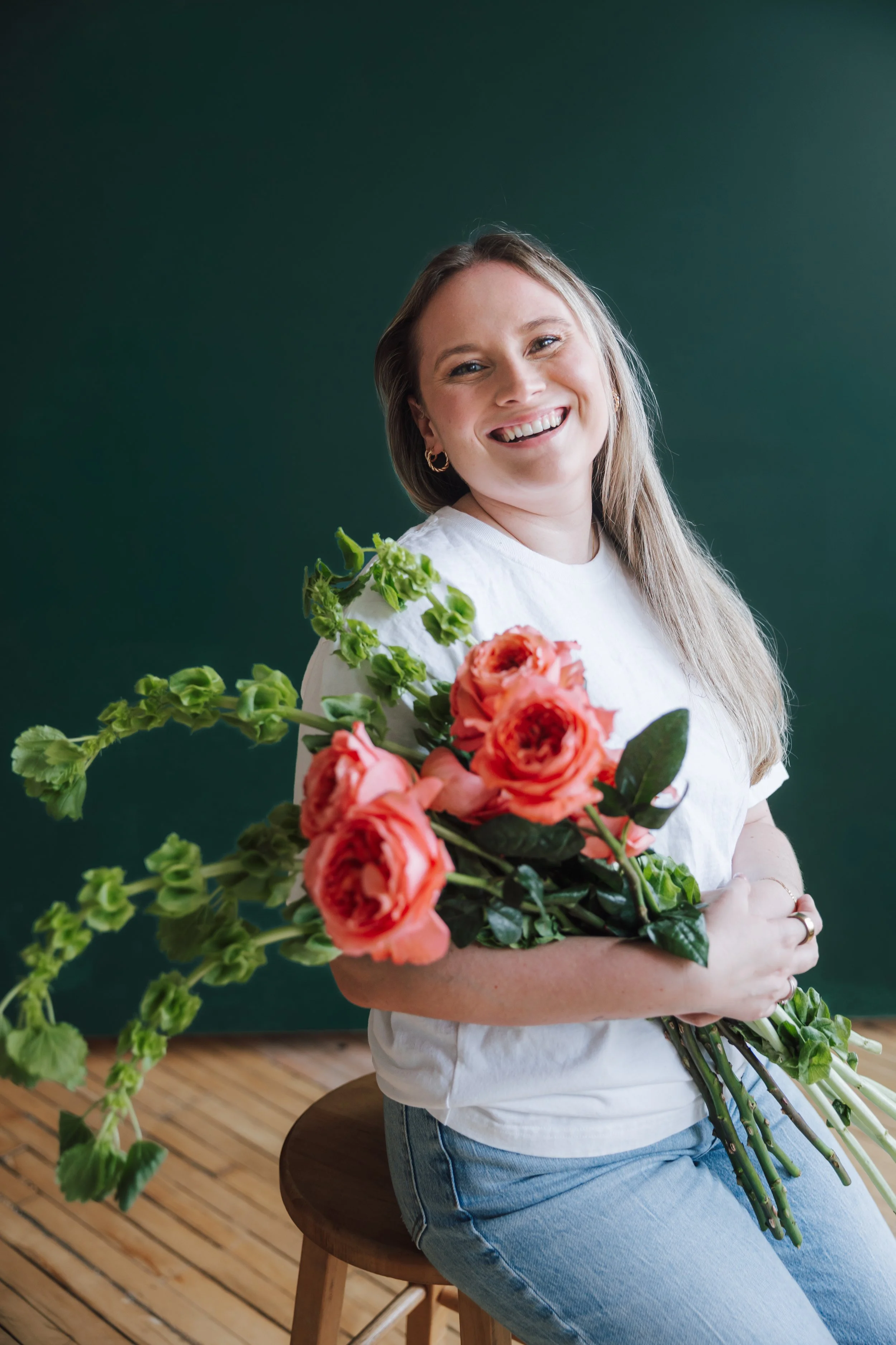 A woman sitting on a wooden stool, smiling, holding a bouquet of orange flowers with green leaves, against a green background, wearing a white t-shirt and blue jeans.