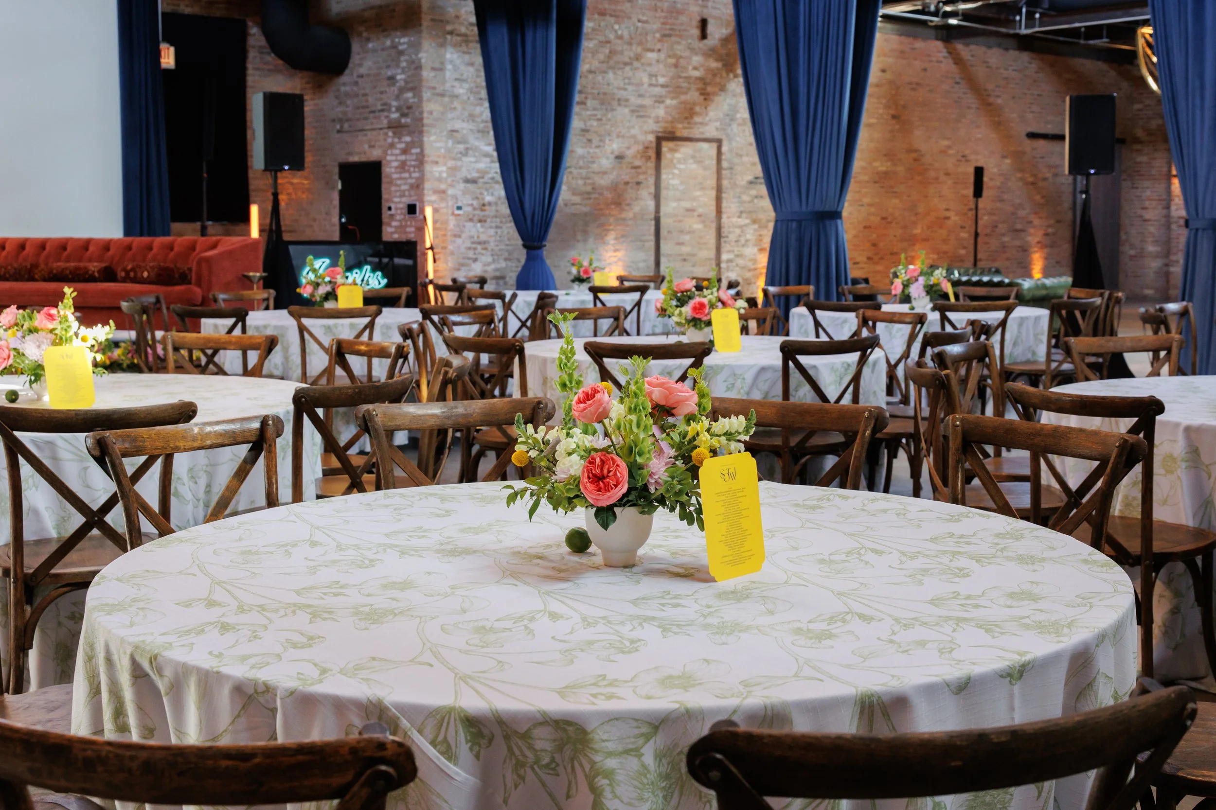 Event space with tables covered in white tablecloths and floral centerpieces, set up for a gathering with a stage in the background, brick walls, and blue curtains.