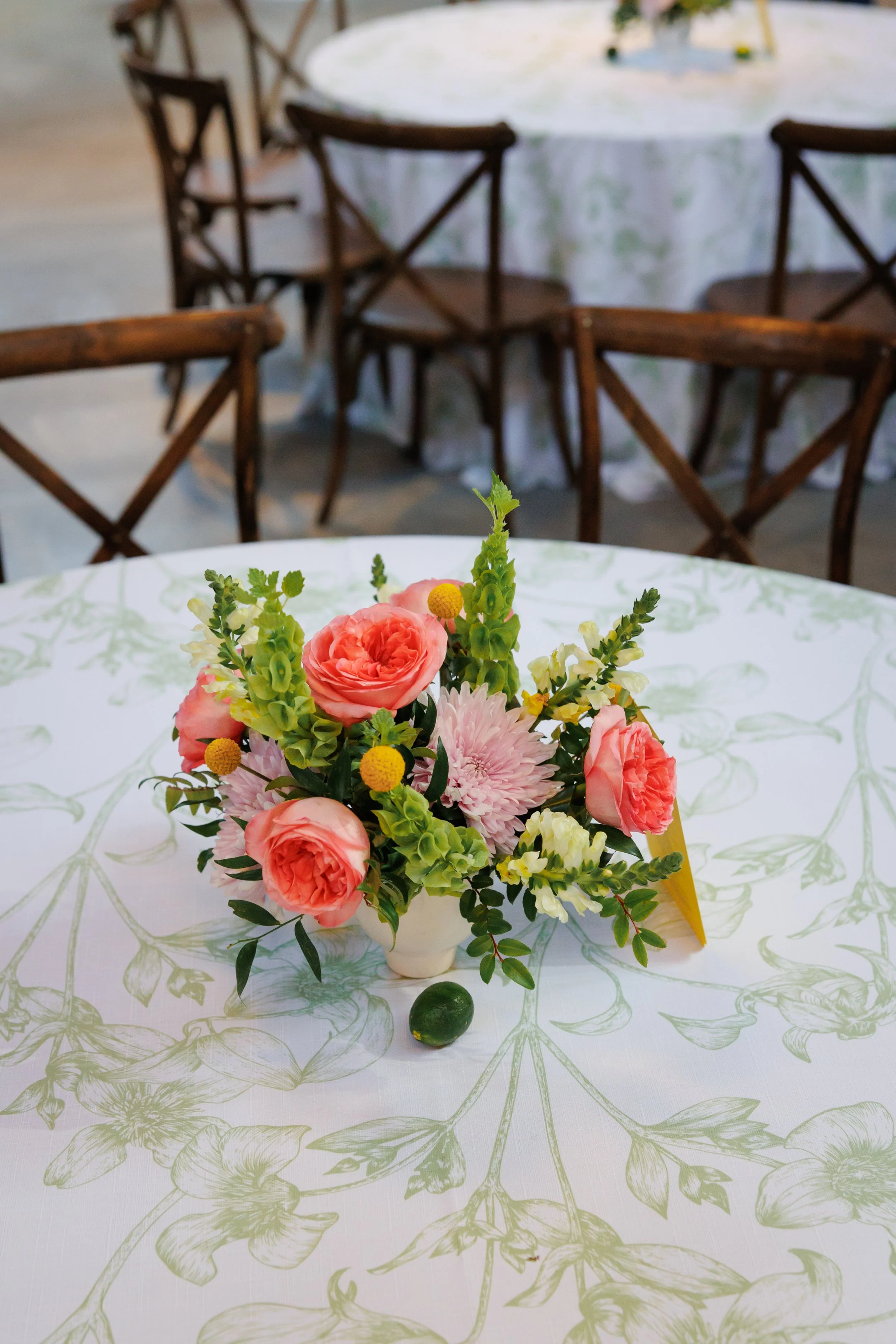 A centerpiece of pink, white, and yellow flowers on a table with a white tablecloth featuring a green floral pattern. In the background, there are several empty wooden chairs around other tables.