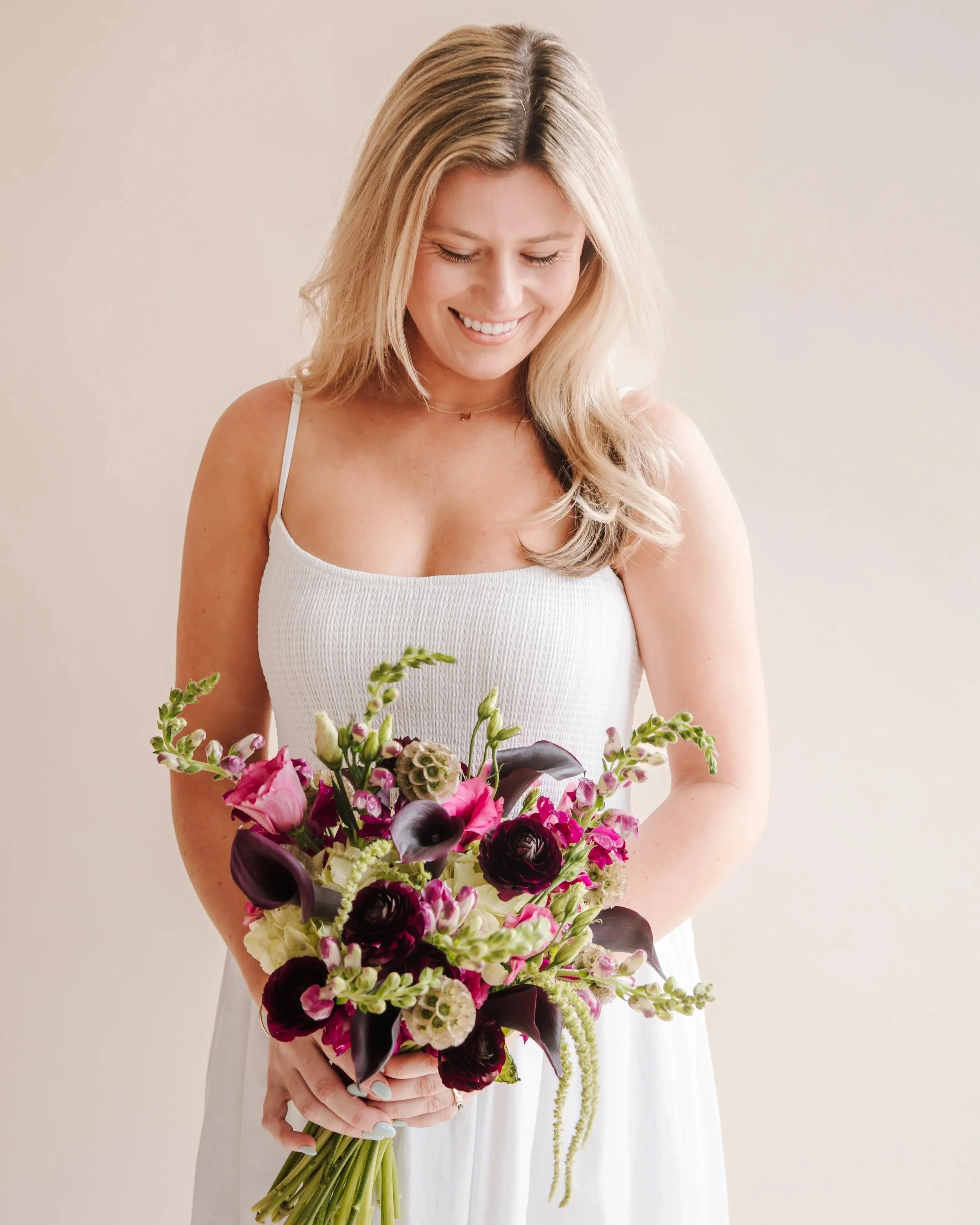 Woman in white dress holding a bouquet of pink and purple flowers, smiling and looking down.