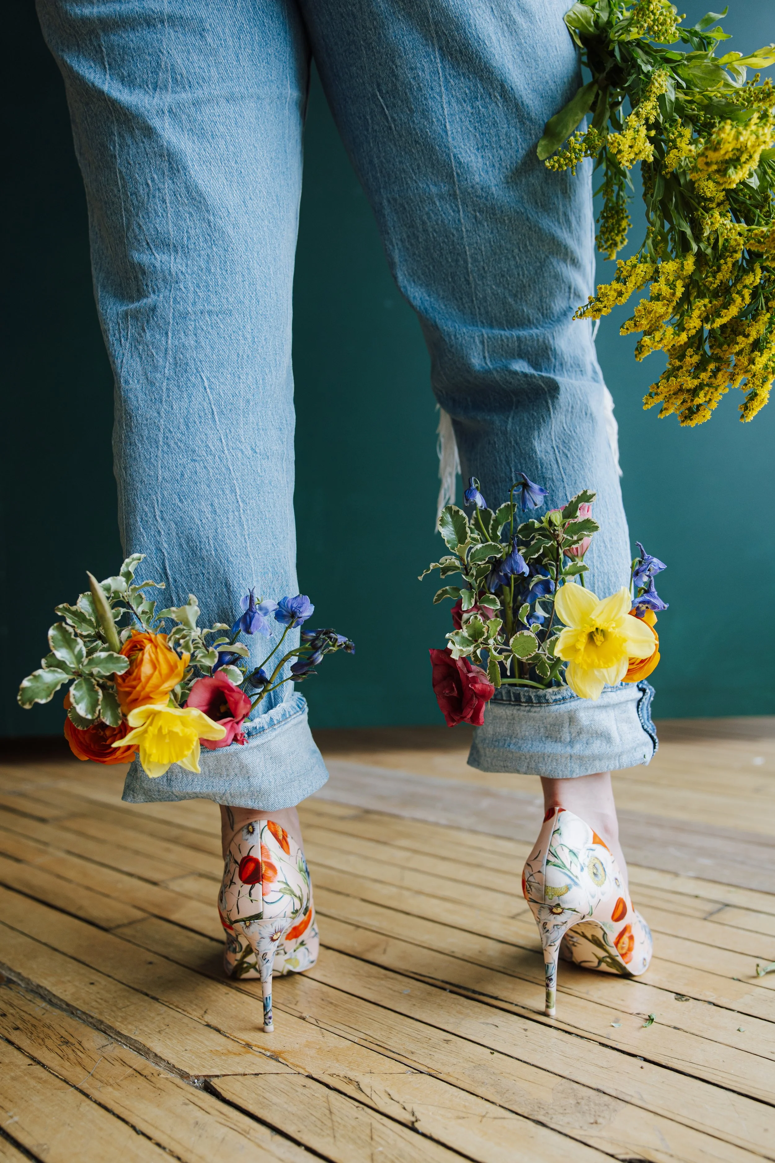 Person wearing floral high heels and blue jeans with the cuffs rolled up, filled with colorful flowers, standing on a wooden floor against a green wall.