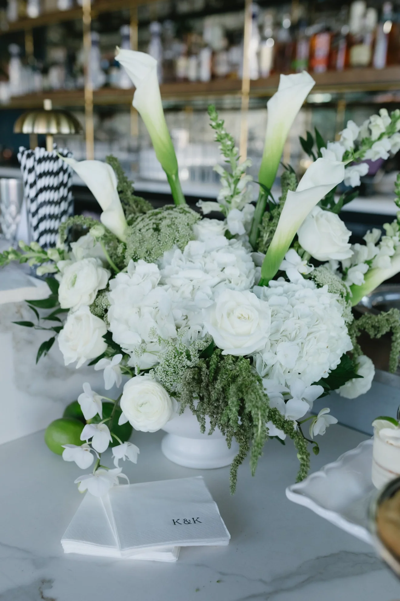 A white floral arrangement with calla lilies, roses, orchids, and greenery in a white vase on a table.