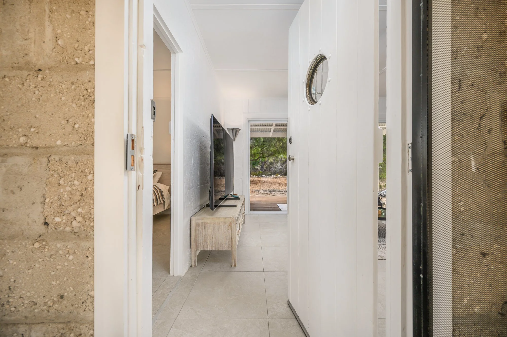 View from between a screen door and a white interior door, looking into a living room with a flat-screen TV on a white stand, a floor lamp, a doorway to a bedroom on the left, and a glass door leading outside to a patio with trees.