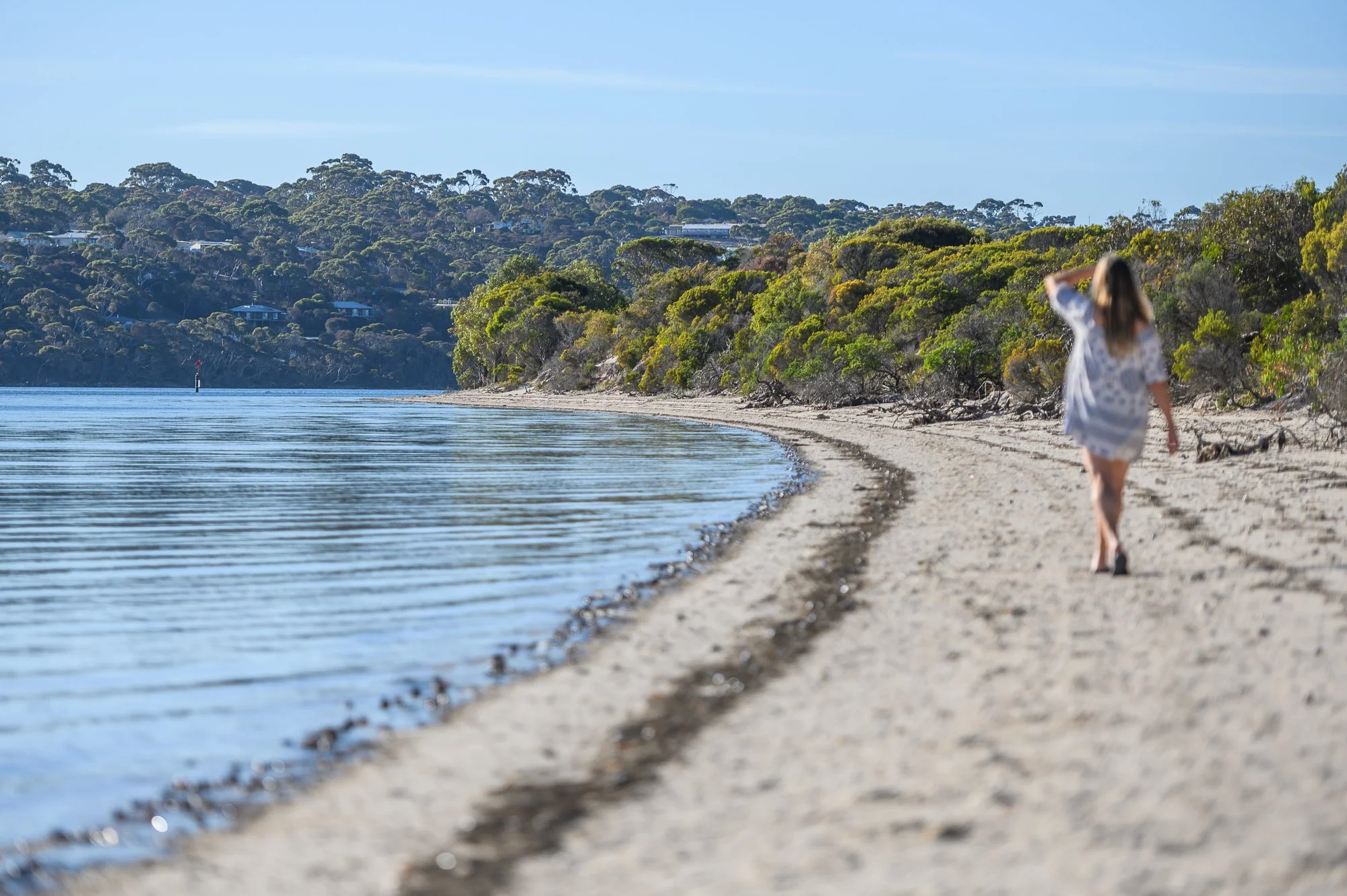 A woman walking along a sandy beach near calm water with trees and hills in the background on a clear day.