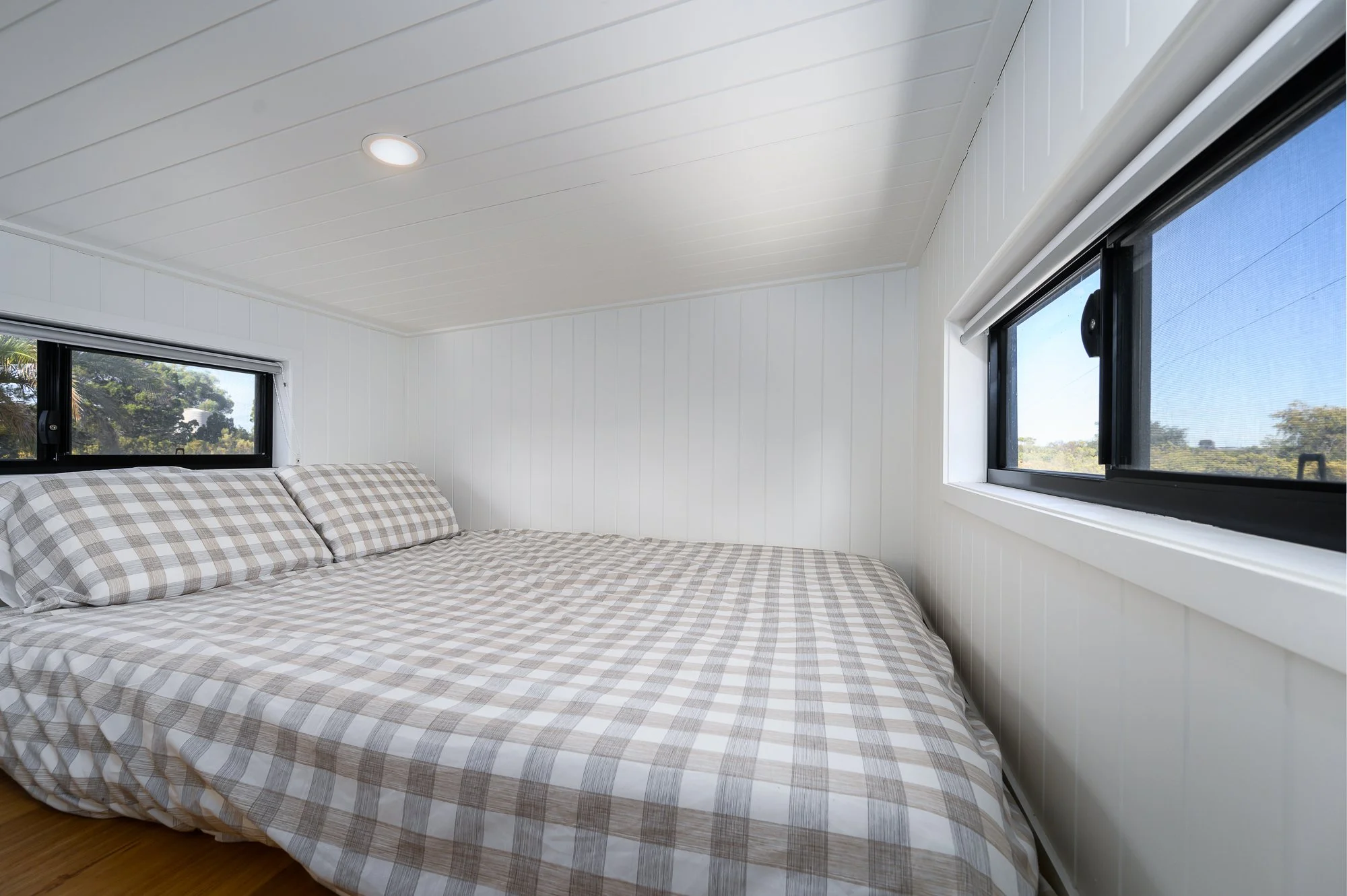Bedroom with a bed covered in checkered bedding, white paneled walls, and two windows showing trees outside.