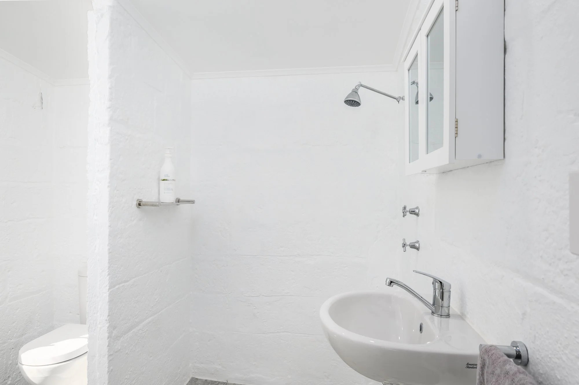 A small, white bathroom with white brick walls, a wall-mounted sink with a chrome faucet, a small mirror cabinet above, a toilet in a separate section, and a showerhead on the ceiling.