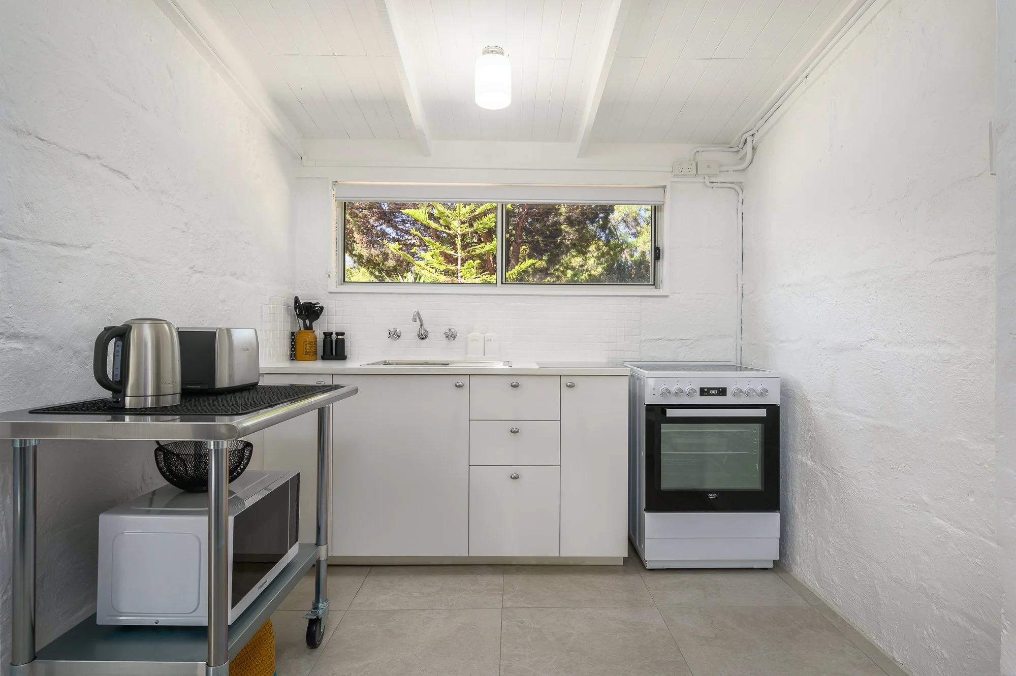 Minimalist kitchen with white cabinets, a window showing outdoor greenery, a stove, a microwave, and kitchen appliances on a trolley.