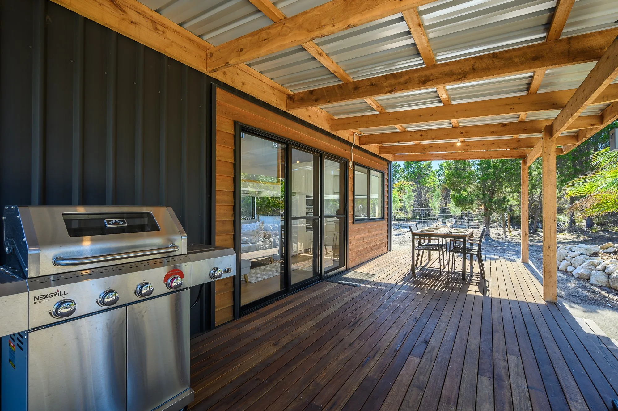 Covered outdoor patio with a stainless steel grill on the left, glass sliding doors leading into a house, a black outdoor dining table with four chairs on the right, and a rural landscape with trees and rocks in the background.