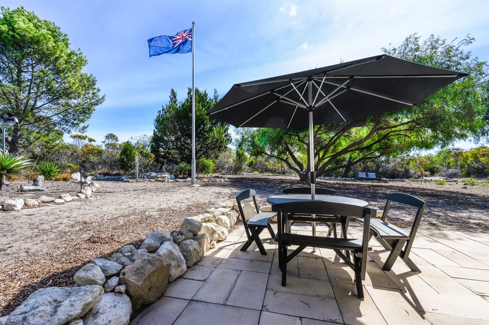 Outdoor patio with a black table, four black chairs, and a large black umbrella on a paved surface. A garden with plants and trees, a flagpole with a flag, and benches in the background under a clear blue sky.