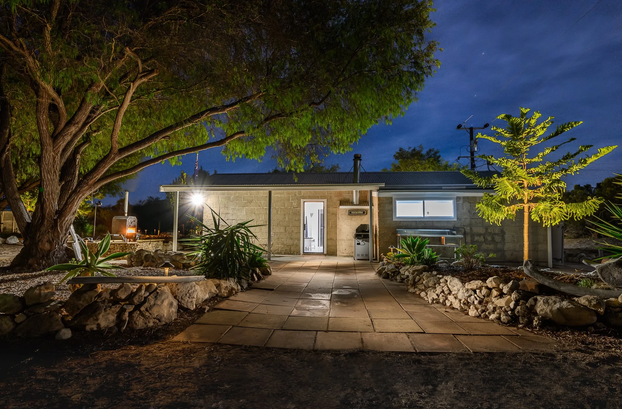 Nighttime view of a small house with stone walls, illuminated by exterior lighting, surrounded by trees, rocks, and desert plants, with a paved walkway leading up to the front door.