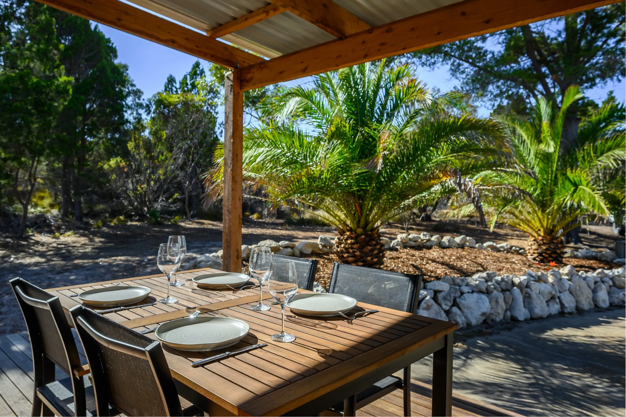 An outdoor dining table set with plates, wine glasses, and utensils on a wooden deck, with a backdrop of palm trees and lush greenery.