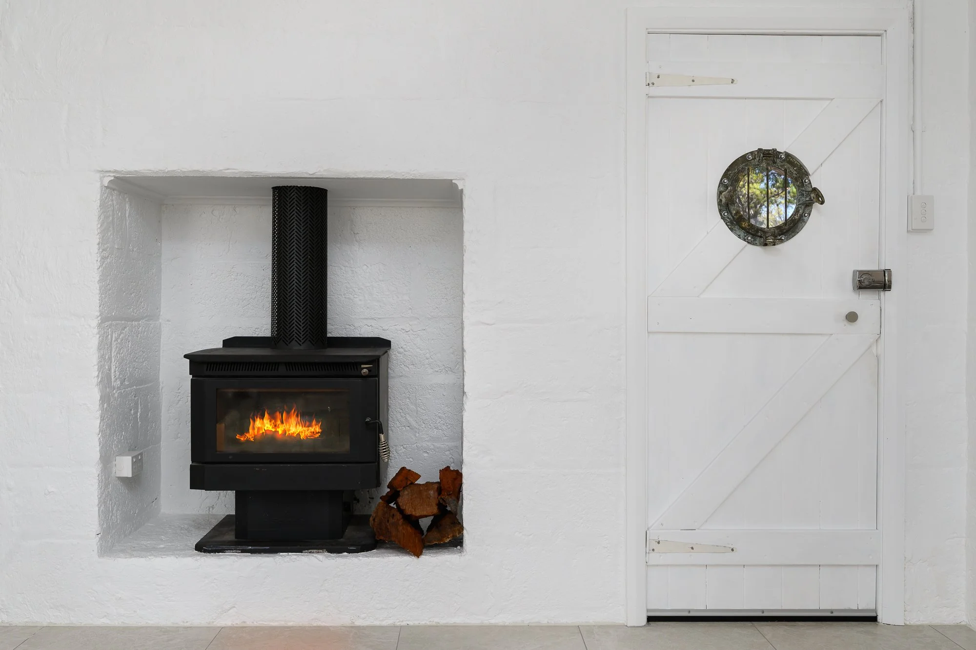 A cozy white room with a black wood stove burning, with logs stacked beside it, next to a white wooden door with a round porthole window and a latch.