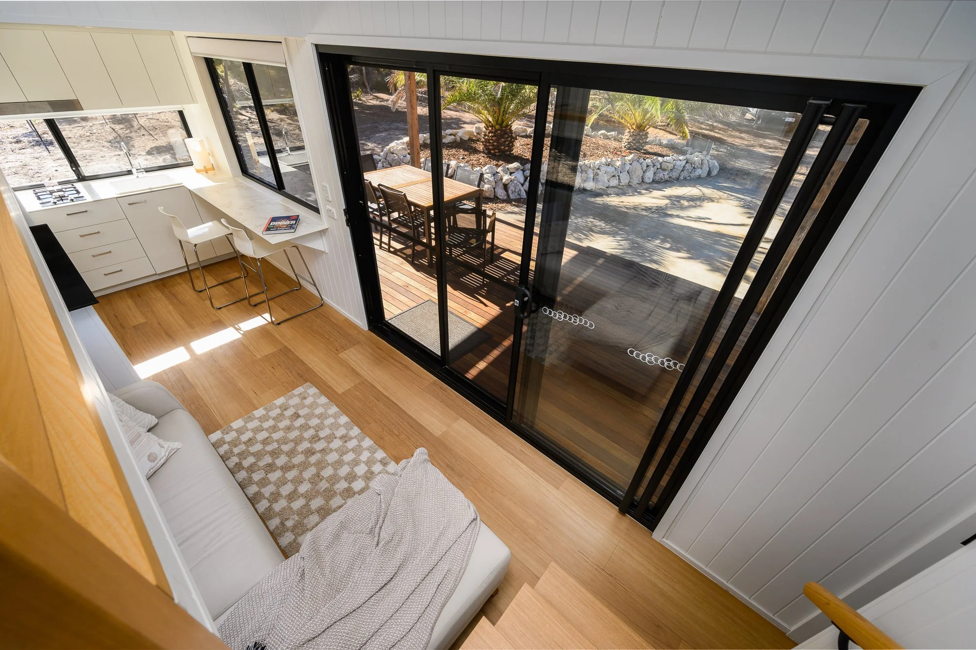Interior view of a modern kitchen and living room with a sliding glass door leading to an outdoor patio with a table and chairs, and a landscaped yard with rocks and palm trees.