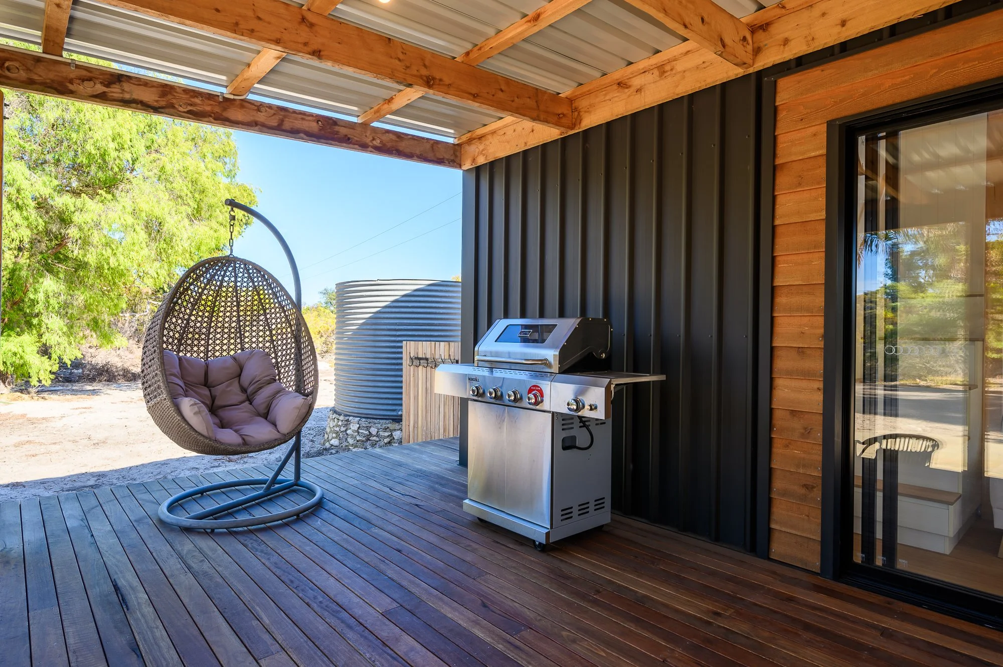 Outdoor patio with wooden flooring, a hanging egg-shaped wicker chair with cushions, a stainless steel gas grill, a large metal water tank, and a sliding glass door leading inside, with greenery and trees in the background.