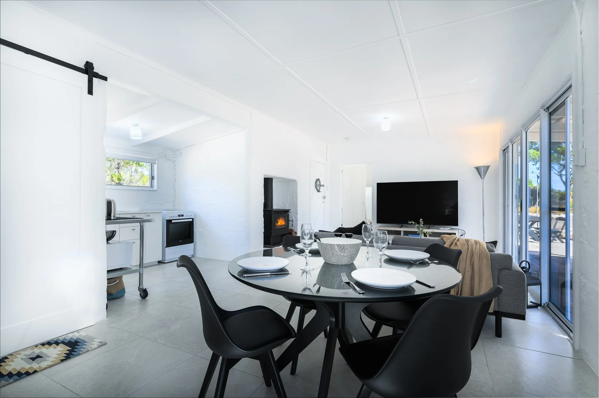 Interior of a modern, bright living and dining area with white walls and ceiling, a black dining table with four black chairs, place settings, a large flat-screen TV, a sofa, a fireplace, and sliding glass doors leading outside.