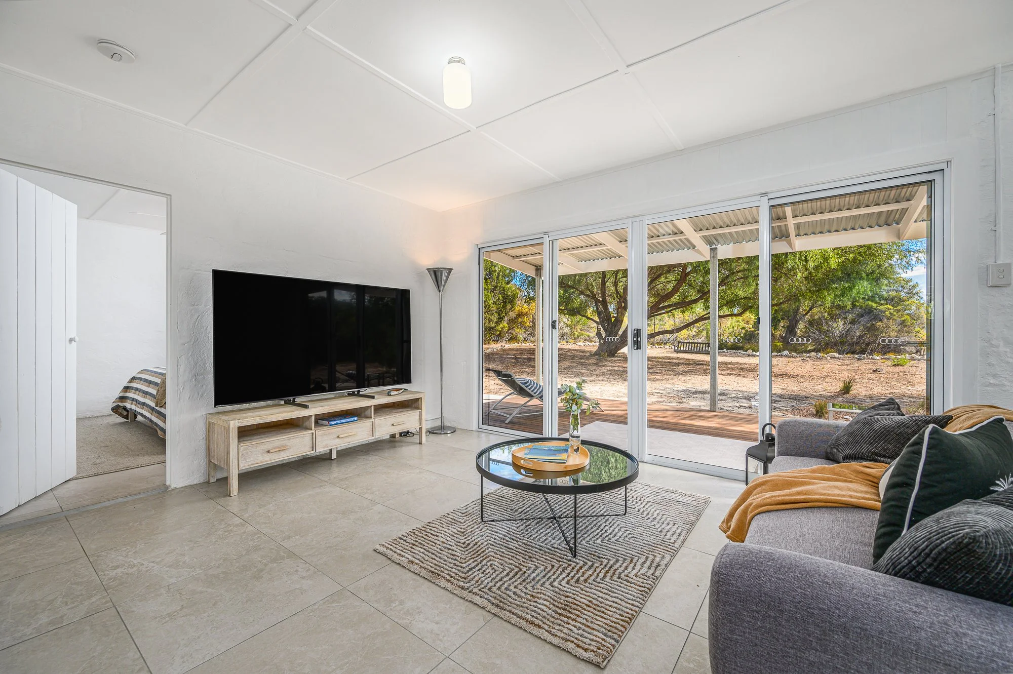 Living room with large sliding glass doors opening to outdoor patio with trees. Contains a gray sofa with pillows, a round black coffee table, a TV on a wooden stand, a floor lamp, and a striped rug.