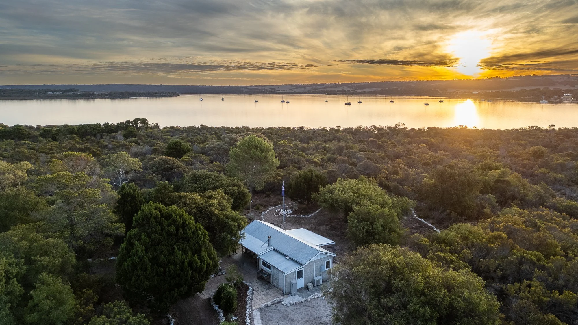 A small house near a lake surrounded by lush green trees at sunset, with boats on the water in the distance.